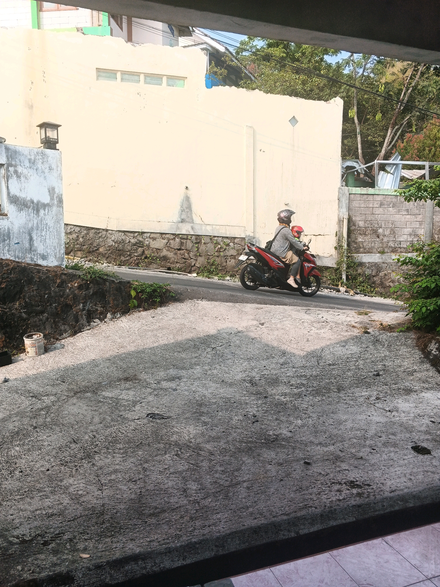 In Keji, Indonesia, on a sloped, paved area, two individuals are riding a red motorcycle. The person in front, wearing a grey shirt and a brown helmet, is operating the motorcycle. The passenger behind them is wearing a red helmet. The motorcycle is moving uphill on a paved road that curves to the right. A light-colored building with a stone foundation is visible behind the road. To the left of the road, a dark, rough-textured wall is present. In the foreground, the paved surface is cracked and textured, with shadows cast across it. Lush green foliage is visible on the right side of the image, and trees with visible power lines are in the background.