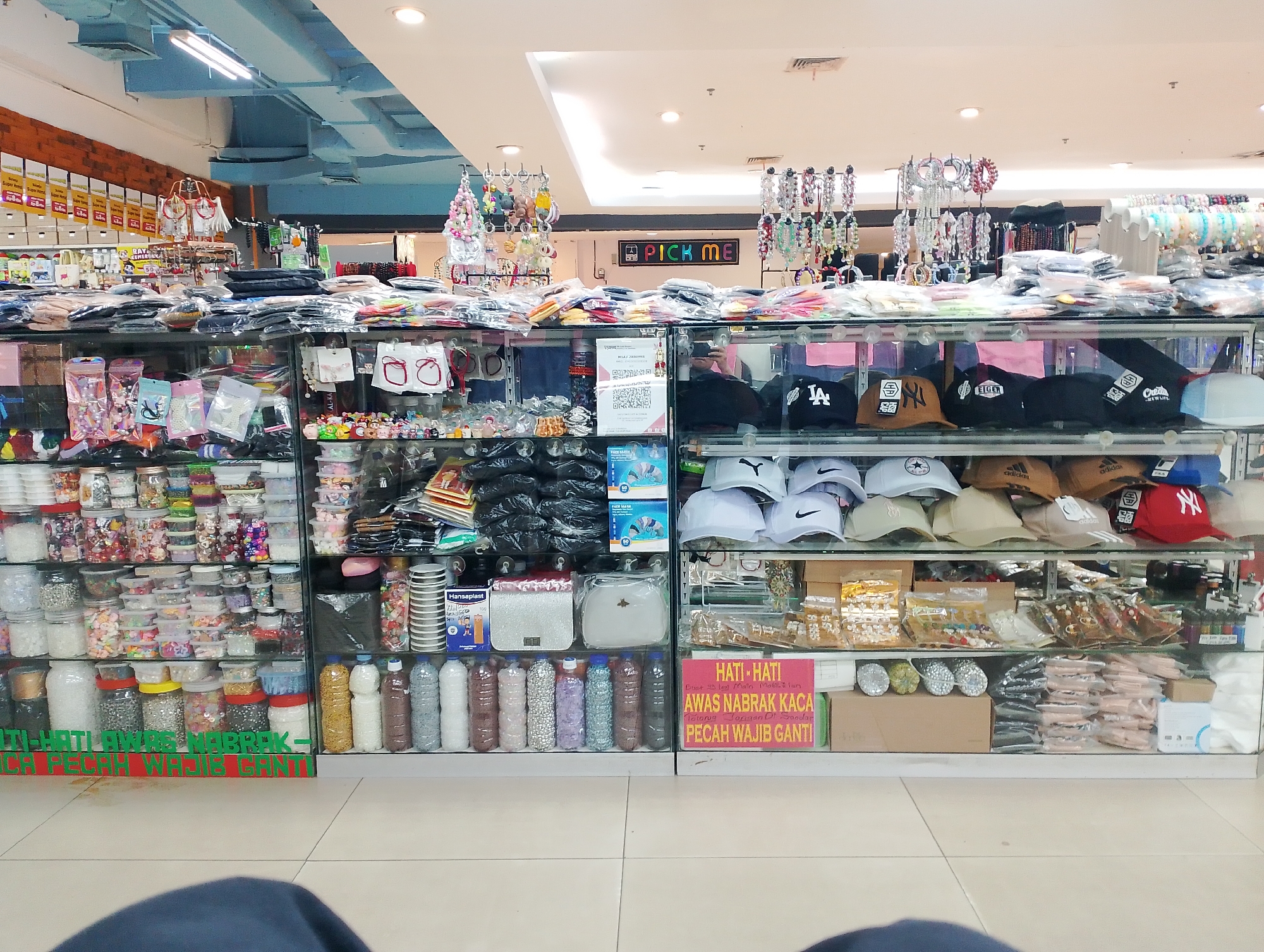 This is a medium-wide shot of a retail display within a store, likely a department store or a large market, in Batam, Indonesia. The image captures a section of a store interior featuring various merchandise displayed on tiered shelves behind glass panels. The lighting appears to be fluorescent, typical of indoor retail environments.
In the foreground, the lower shelves are filled with an assortment of items including what appear to be plastic containers of various sizes and colors filled with small, colorful objects, possibly craft supplies or decorative items. There are also several bottles with what looks like granular material inside. On the middle shelf, there are more packaged items, some in clear plastic bags, and also some handbags or purses. A significant portion of the middle and upper shelves are dedicated to displaying numerous baseball caps and hats, many of which are black with various logos like "LA," "Adidas," and "Eiger." Other accessories like bracelets are displayed on stands above the shelves.
In the background, the store's ceiling is visible with exposed blue ductwork and fluorescent lighting fixtures. A neon sign with the words "PICK ME" in colorful letters is mounted on the wall. Promotional banners and signage are also visible in the upper left portion of the image. The overall impression is one of a well-stocked retail space offering a variety of goods. No people are clearly visible, though reflections in the glass suggest the presence of a photographer or shopper. The floor is tiled, and the overall environment appears clean and organized for retail.