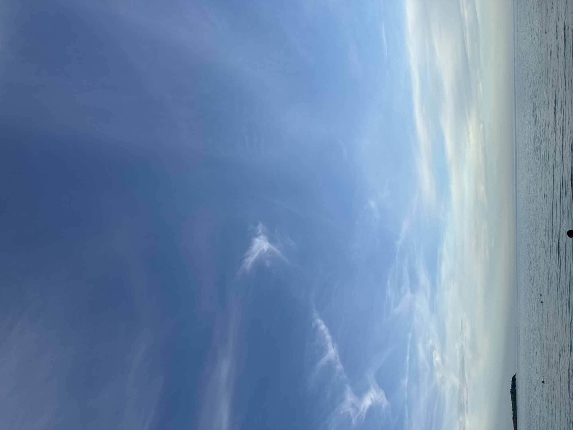 The image shows a serene seascape with a vast expanse of calm water under a blue sky dotted with wispy white clouds. In the distance, a small, tree-covered island is visible on the left. The water's surface reflects the sky, creating a soft, muted color palette. A few indistinct figures can be seen wading in the water near the shore, suggesting a peaceful beach scene. The overall impression is one of tranquility and natural beauty.