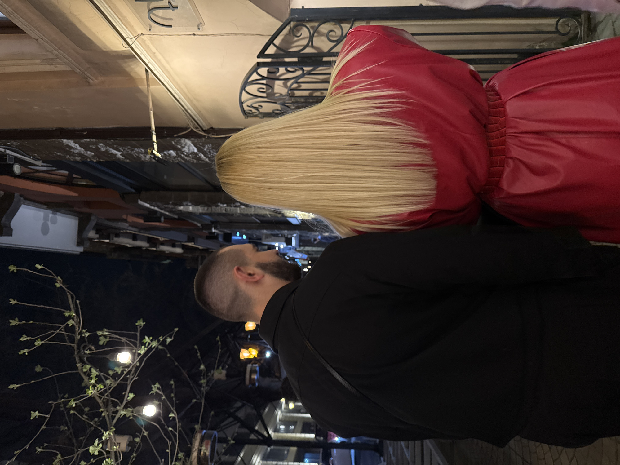 The image captures a nighttime scene in Yerevan, Armenia, based on the architectural style and street lighting. A man with a shaved head and beard is wearing a black shirt with a strap visible across his back. He stands beside a woman with long, straight blonde hair, who wears a red dress that appears to be made of leather or a similar material. They appear to be looking up at something unseen. The surroundings include buildings with visible windows, street lamps, and trees with small leaves, which suggests it may be spring or early summer. There is an ornate metal railing or gate behind the woman.