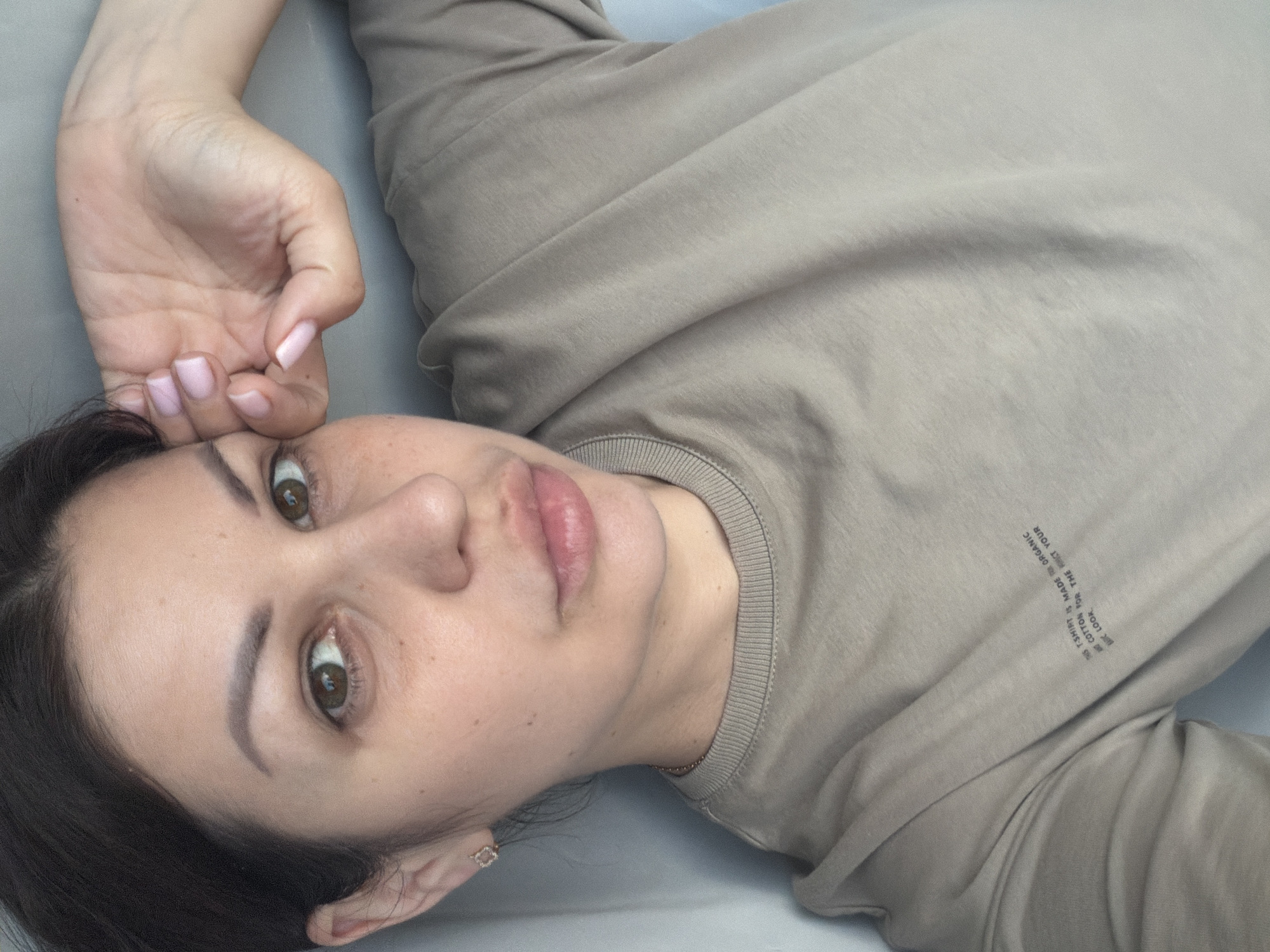 A woman is lying on her back, looking at the camera. She has dark hair, brown eyes, and light skin. She is wearing a tan t-shirt with a small print on the left side of the chest that reads "THIS T-SHIRT IS MADE FROM ORGANIC COTTON FOR THE MINIMAL LOOK." Her left hand is raised to her face, with her fingers touching her forehead. Her nails are painted a light pink. She is wearing a small, square earring in her left ear. The background is a plain, light-colored surface, possibly a wall or a bed. There are no identifiable landmarks or contextual clues in the image to suggest a specific location within the city of Sochi, Russia.