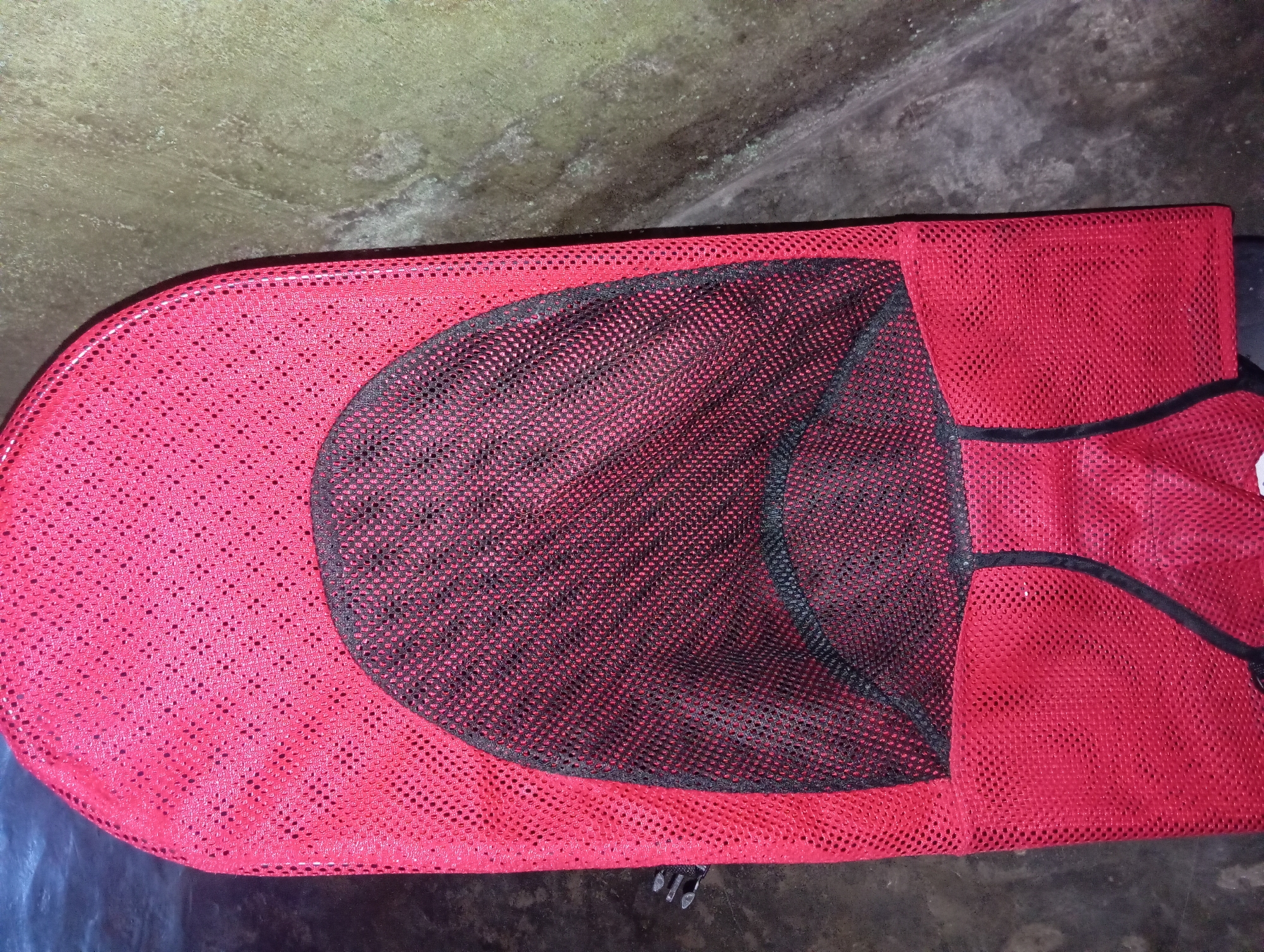 A close-up, overhead shot shows a red mesh baby bouncer with a black mesh insert. The bouncer is positioned on a mottled gray surface, possibly concrete, with a dark, textured background to the left. The bouncer has a rounded top and tapers towards the bottom, with black trim along the edges and a black seam running down the center. The mesh material of the bouncer is visible, with small, evenly spaced holes. The lighting appears to be artificial, casting slight shadows. No people are present in the image. The overall impression is one of an indoor setting, possibly a home, as the surface does not appear to be outdoors. The specific location in Rejosari, Indonesia, cannot be determined from the visual information provided.