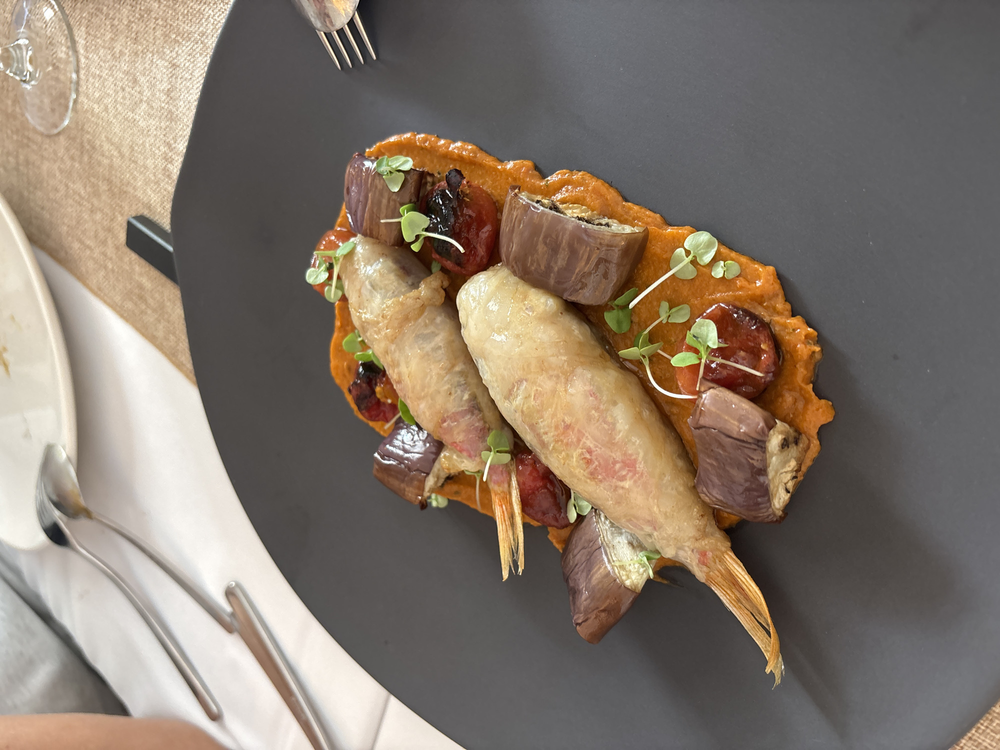 An overhead shot of a gourmet dish, likely taken at a restaurant in Carboneras, Spain, showcases two whole fish, lightly battered and fried, resting on a vibrant orange puree. The fish are adorned with small sprigs of microgreens. Surrounding the fish are pieces of roasted eggplant and what appear to be cherry tomatoes, adding color and texture to the presentation. The dish is served on a dark gray, irregular-shaped plate, set on a table covered with a neutral-toned placemat. In the background, silverware and a partially visible wine glass suggest a dining setting. A person's shoulder and arm are visible in the upper left corner, indicating their presence at the table.