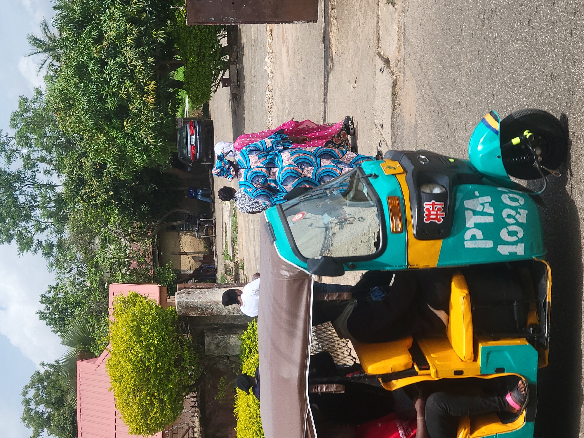 The image captures a bright, sunny day scene on a street in Jos, Nigeria. An auto rickshaw, painted in bright turquoise and yellow, is parked on the side of the road. The vehicle is labeled "PTA-1020." Several passengers are seated inside. Two women are walking past the auto rickshaw; one is wearing a blue and white patterned gown with a matching headscarf, while the other is wearing a pink patterned gown. A black car is visible parked further down the street, under a tree. In the background is vegetation and buildings, typical of an urban environment.