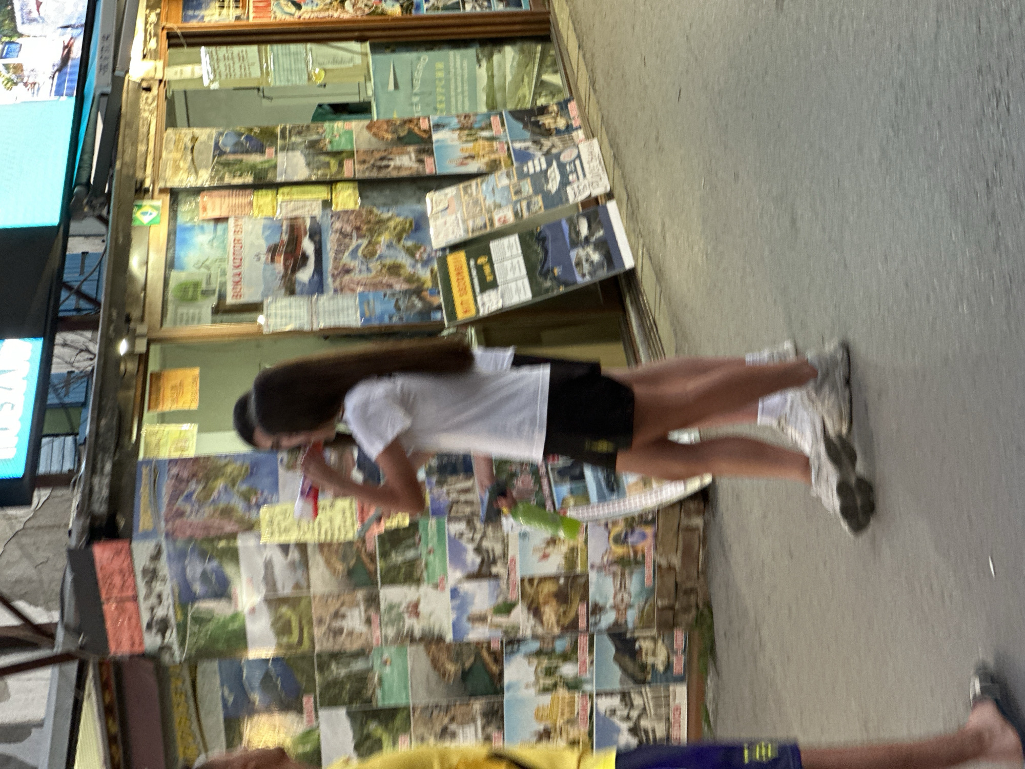 In Budva, Montenegro, two young individuals are captured in a motion blur as they walk along a paved street. One, a girl with long dark hair, is wearing a white t-shirt and black shorts, holding a green bottle and what appears to be a red and white toy. The other, a girl with dark hair, is partially visible, dressed in a white t-shirt and black shorts. To their left, a man in a yellow t-shirt and blue shorts is also visible. Behind them, a storefront displays numerous travel posters and brochures, suggesting a tourist-oriented area. The posters feature various scenic locations, including what appears to be the Bay of Kotor. The storefront itself has large glass windows, and above it, a blue neon sign is visible. The overall atmosphere suggests a lively street scene in a popular tourist destination.