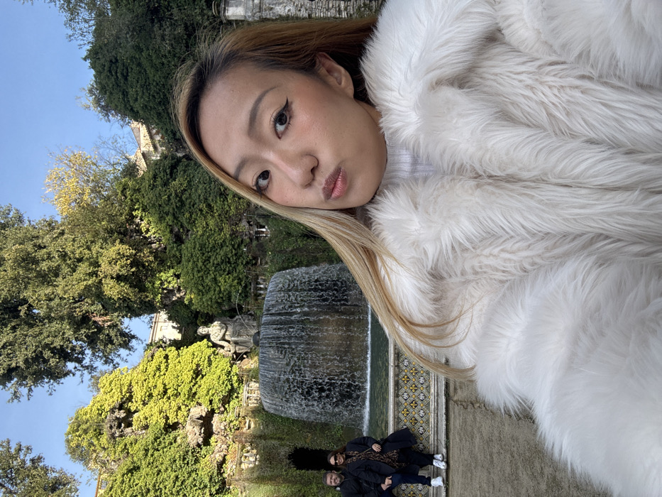 A selfie-style portrait captures a woman in the foreground against the backdrop of the elaborate gardens and fountains of Villa d'Este in Tivoli, Italy. The woman, positioned on the right and center, has long, light brown hair, visible makeup including winged eyeliner, and a slight pout. She wears a thick, shaggy white faux fur coat over a white turtleneck.
In the midground, a large, multi-tiered fountain dominates the scene, featuring a wide, semi-circular cascade of water falling into a lower basin. Above the waterfall, a classical stone statue of a seated figure is visible, surrounded by lush green foliage. Below the fountain, two additional individuals are seated on a decorative tiled ledge with a yellow, blue, and white geometric pattern. The person on the left is a man with a beard, wearing a dark jacket and white sneakers. Next to him, a woman wears dark sunglasses, a dark coat, and a patterned scarf.
The background consists of densely vegetated terraced gardens with various shades of green trees and bushes, some covered in ivy. Portions of a light-colored building are visible behind the upper garden tiers. The sky is clear and blue, indicating bright daylight conditions.