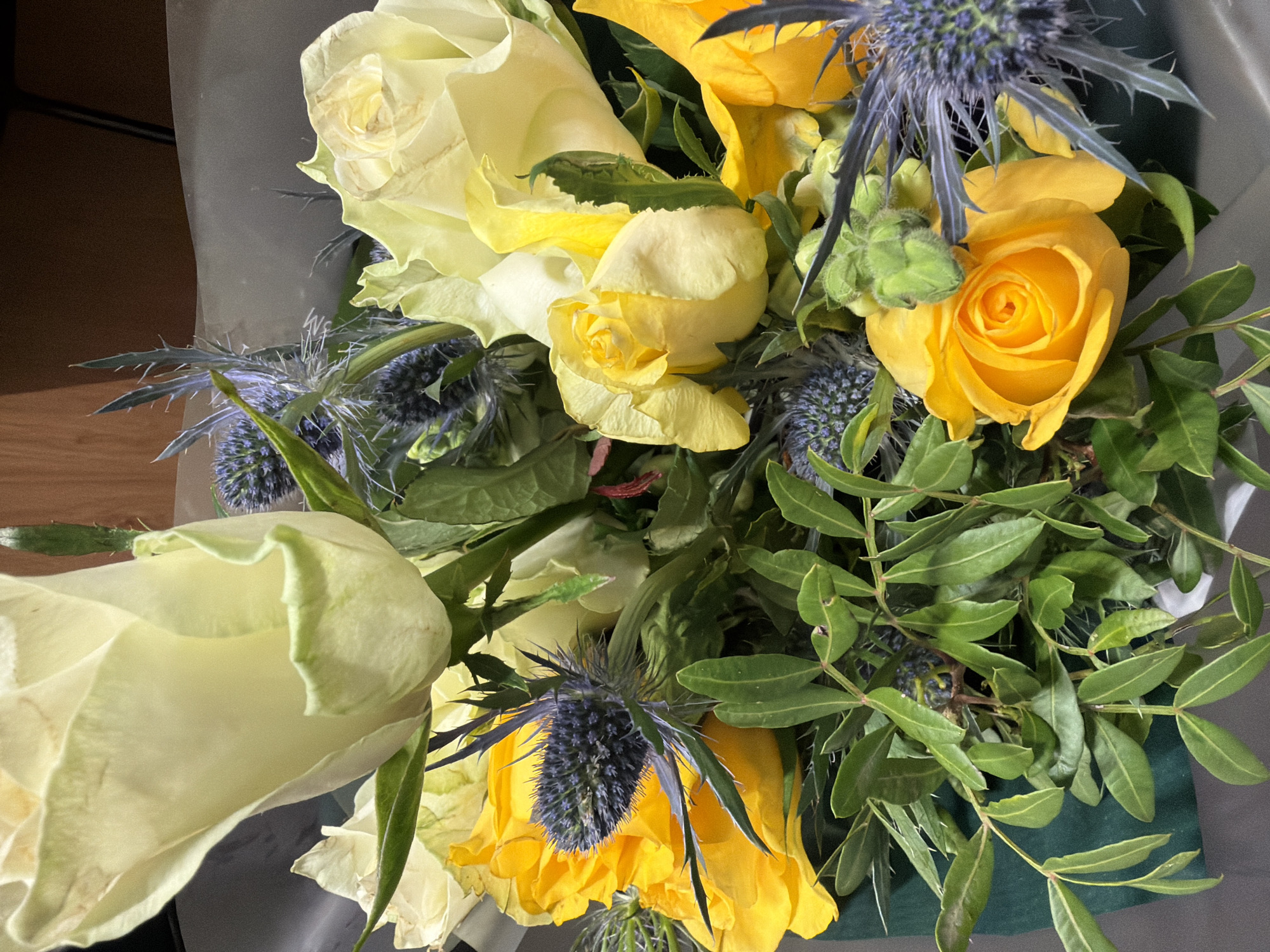A close-up shot depicts a bouquet of flowers, primarily composed of yellow and white roses, interspersed with blue thistle-like flowers and green foliage. The scene is focused on the floral arrangement itself, with the background partially visible and consisting of what appears to be wrapping paper or packaging. The colors are vibrant, with the yellow and white roses providing a contrast to the darker blue and green elements. The composition is dense, with the flowers and foliage closely packed together. There is no indication of a specific location in London, United Kingdom, nor any people or interactions evident in the image.