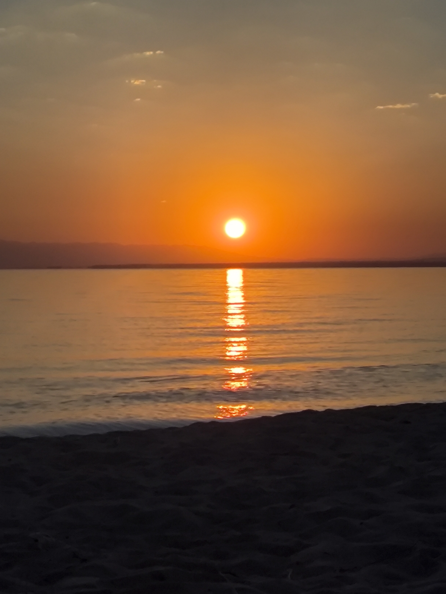The image captures a serene sunset over a body of water, likely a lake or sea, as viewed from a sandy beach. The sun, a brilliant orange orb, is low on the horizon, casting a warm, golden glow across the sky and reflecting a shimmering path onto the water's surface. The sky is a gradient of orange and pale blue, with a few wispy clouds scattered across it. In the distance, a dark, silhouetted landmass is visible, hinting at distant shores or mountains. The foreground is dominated by the dark, textured sand of the beach, which is slightly out of focus, suggesting a close-up perspective of the shoreline. The overall impression is one of tranquility and natural beauty.