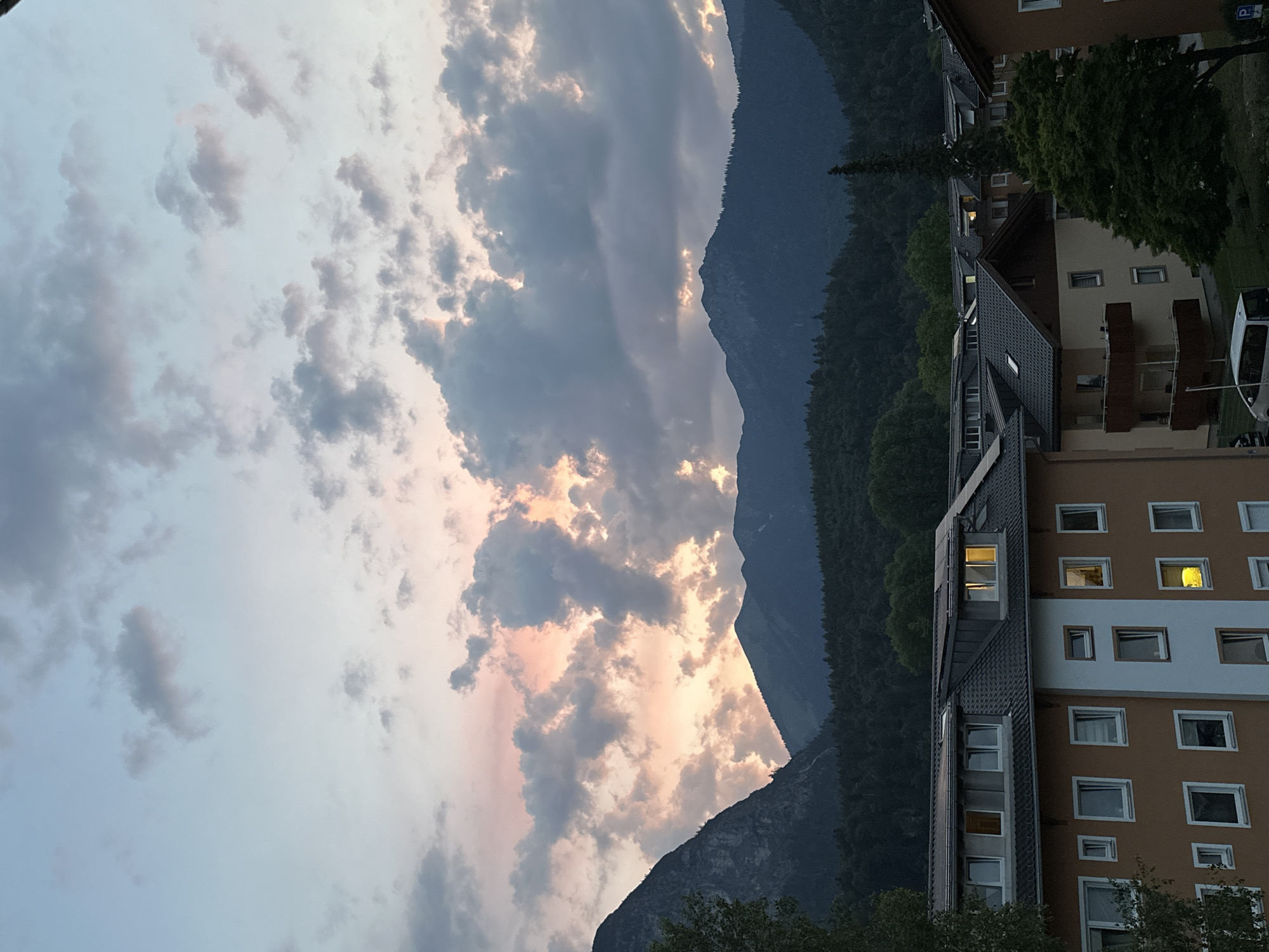 The image depicts a twilight scene in Garmisch-Partenkirchen, Germany, with the Alps as a prominent backdrop. The sky is filled with dramatic clouds, tinged with soft pinks and oranges from the setting sun, contrasting with the darkening blue and gray hues. In the foreground, several multi-story residential buildings with light brown and white facades are visible. Their windows are mostly dark, but a few emit a warm, yellow glow, suggesting indoor lighting. The buildings have dark, sloped roofs, some equipped with solar panels. A dense forest covers the lower slopes of the mountains, transitioning into the rugged, dark peaks that dominate the horizon. The overall atmosphere is serene and tranquil, characteristic of a European alpine town at dusk.