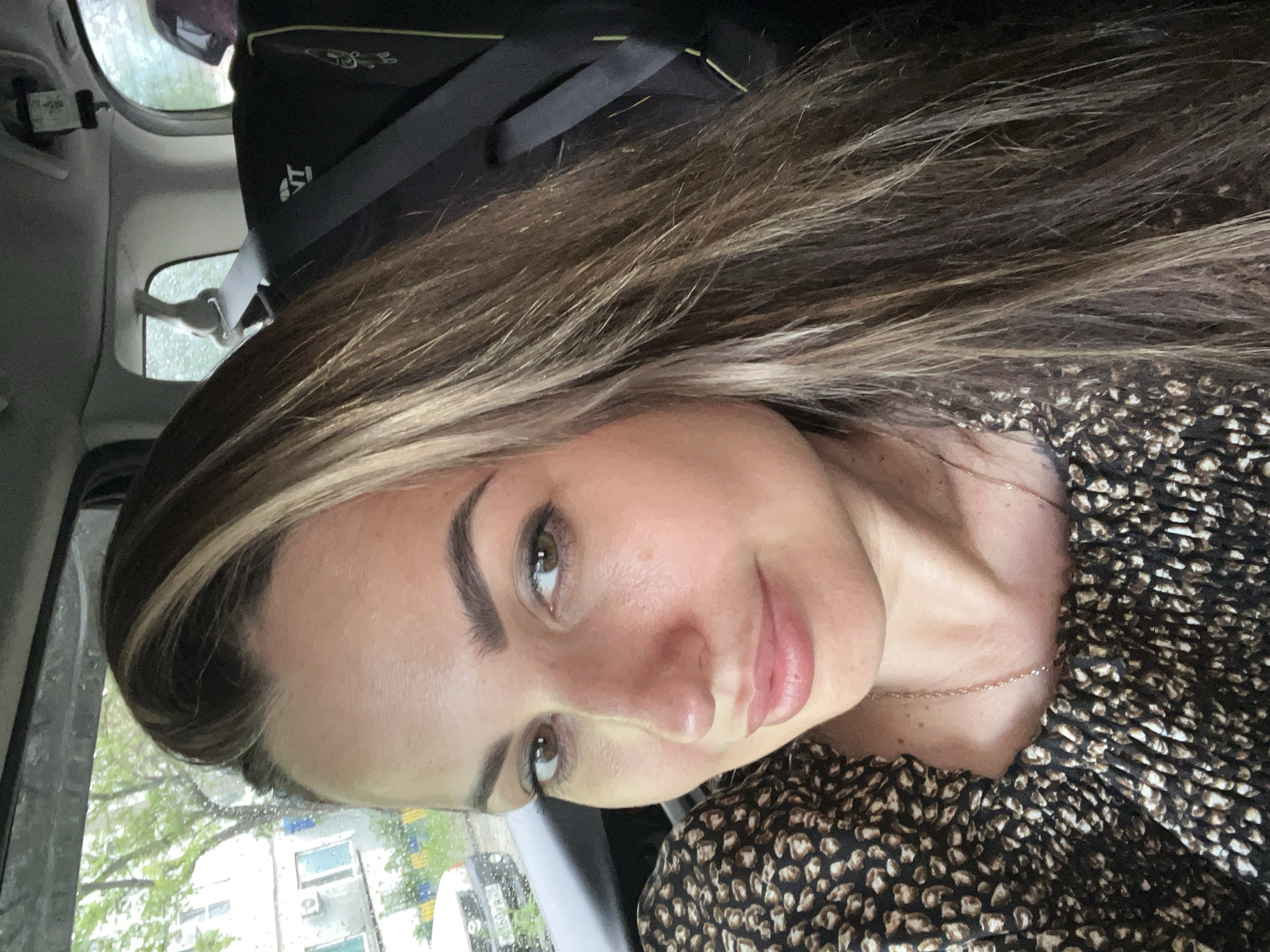 A medium shot shows a woman in the front passenger seat of a vehicle, looking towards the camera. She has long, wavy brown hair with blonde highlights, and is wearing a black and brown patterned top. The woman's face is slightly turned to her left, with her eyes looking directly at the viewer. Her makeup is natural, with emphasis on her eyebrows and lips. A delicate gold necklace is visible around her neck. The background shows the interior of a car, with a child car seat visible behind her. Outside the car window, a residential building with trees and a street can be seen, suggesting an urban setting. The weather appears to be overcast.
