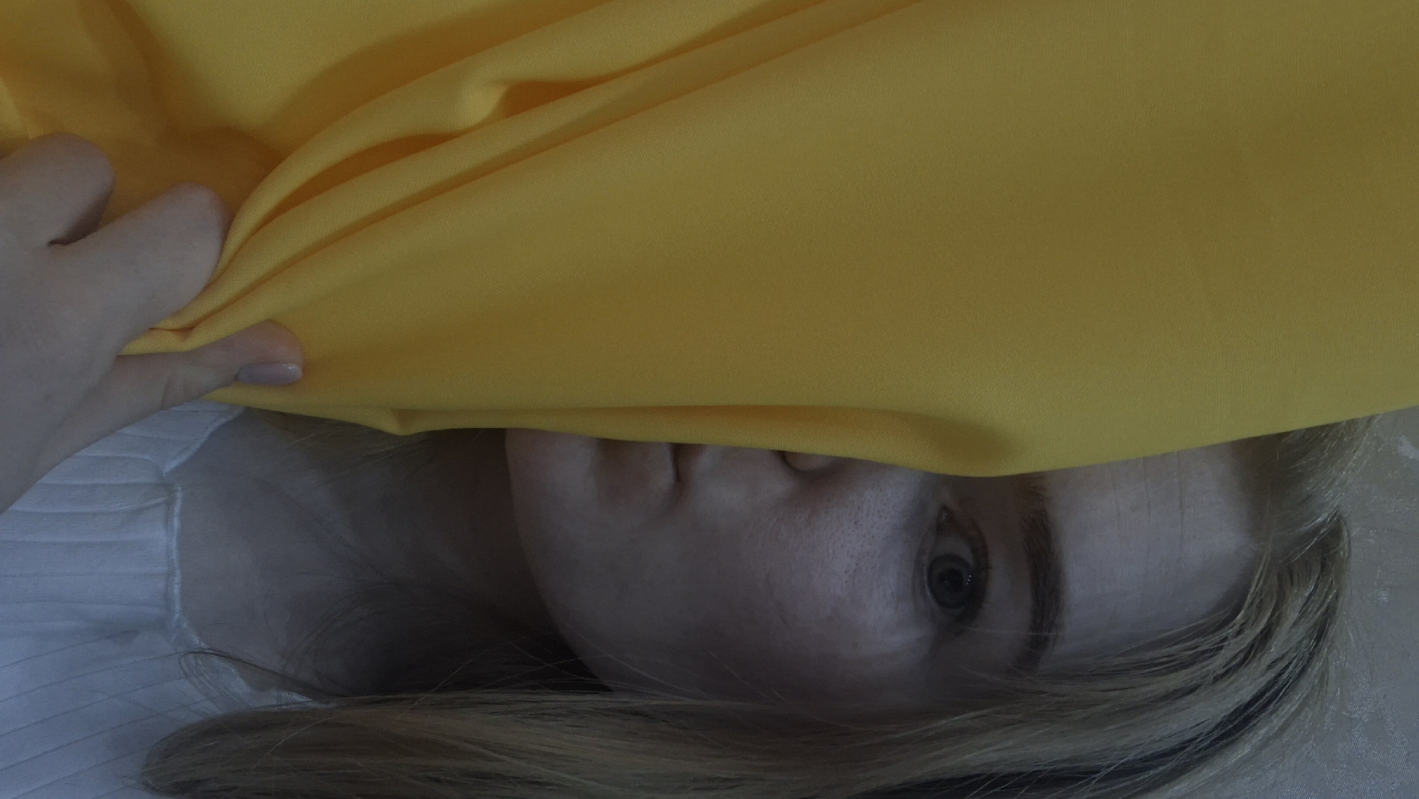 A fair-skinned person with blonde hair is partially visible, lying down. A hand holds up a mustard-yellow fabric, partially obscuring the face. The environment is soft and suggests an indoor setting, potentially on a bed. There is no information available about the location in the city of Chaikovsky, Russia.