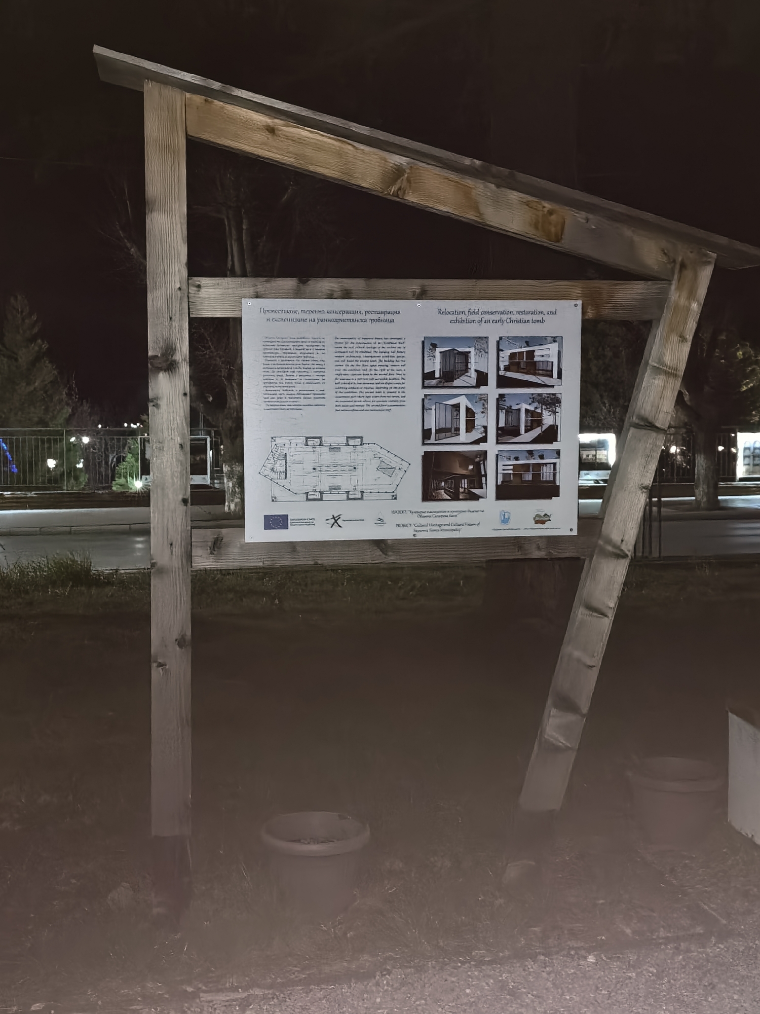Here is a detailed description of the image:
The image shows a wooden sign with information about the "Relocation, field conservation, restoration, and exhibition of an early Christian tomb". The sign is set up near a street, with grass and trees visible in the background. The photo was taken at night, and the surroundings are dimly lit. The sign has a title in both Bulgarian and English, with text explaining the project. Below the text, there are pictures illustrating different stages of the project and the logo of "Cultural Heritage and Local Identity". Beside the wooden sign, there is an outdoor pot.