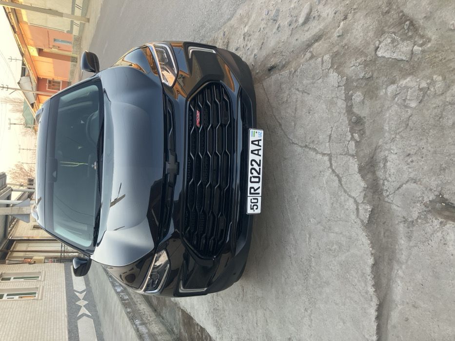 The image displays the front of a black Chevrolet Tracker SUV, parked on a cracked concrete surface in a residential area of Sang, Uzbekistan. The vehicle features a blacked-out Chevrolet bowtie emblem and a red "RS" badge on its dark, hexagonal grille. The LED daytime running lights are visible. A white Uzbekistan license plate, bearing the alphanumeric sequence "50 R 022 AA", is mounted on the lower front bumper. A green-beaded ornament hangs from the rearview mirror inside the vehicle. To the left, a light-colored brick building with a distinctive gray chevron pattern along its lower section is visible. The background shows a street lined with residential buildings of varying colors including beige, terracotta, and light orange. Overhead utility lines and sparse, bare trees are present further down the street. No people are visible, and no actions are in progress. The lighting suggests a clear day.