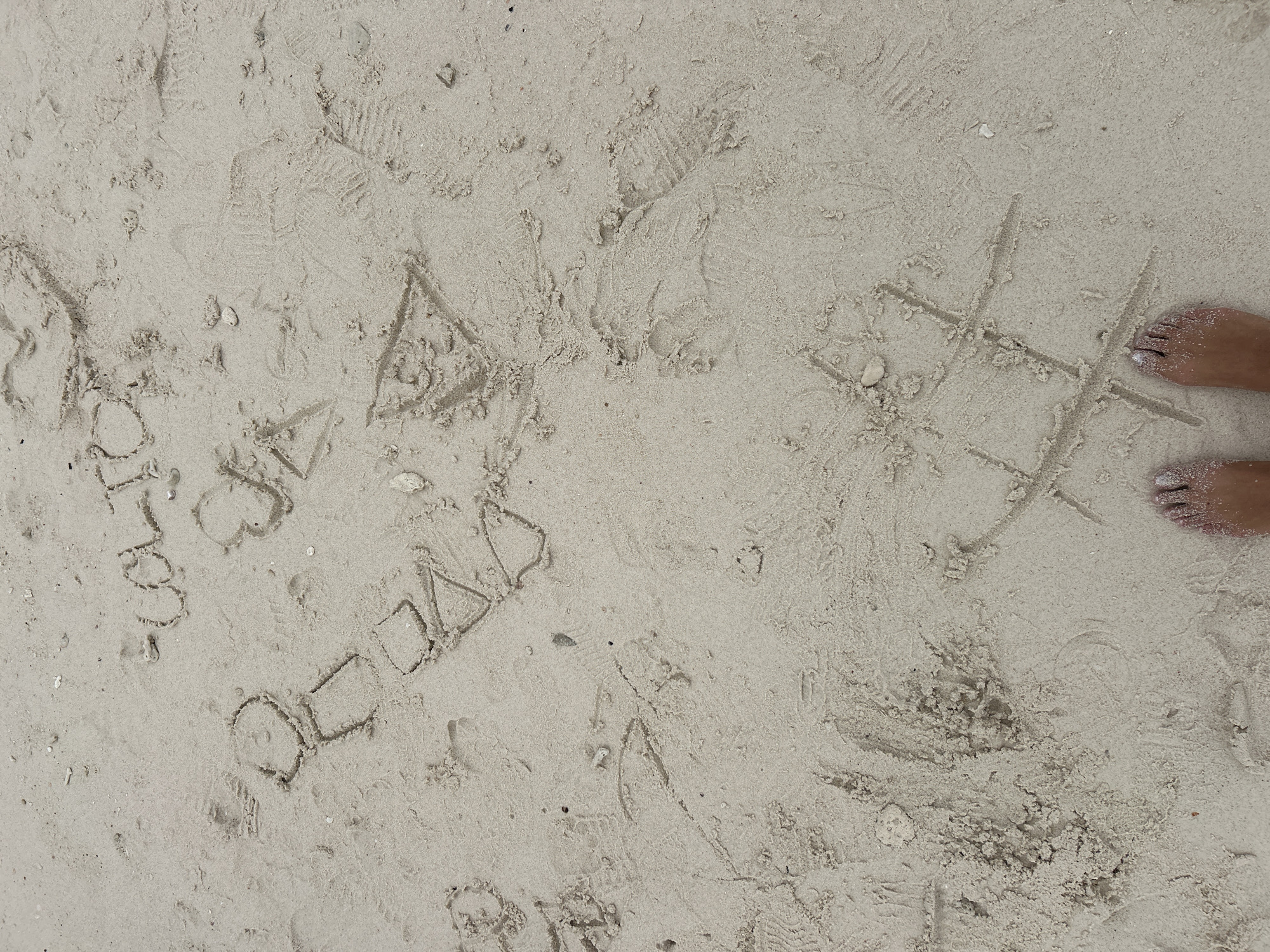 In Phru Nai, Thailand, on a sandy beach, a game of tic-tac-toe is drawn in the sand. Nearby, the words "CAIO" and "ROSA" are also etched into the sand. Two pairs of feet, one wearing nail polish, are visible at the bottom of the frame, indicating the presence of people. The sand appears damp in some areas, suggesting recent activity or proximity to the water. Footprints are visible throughout the sand, indicating foot traffic in the area.