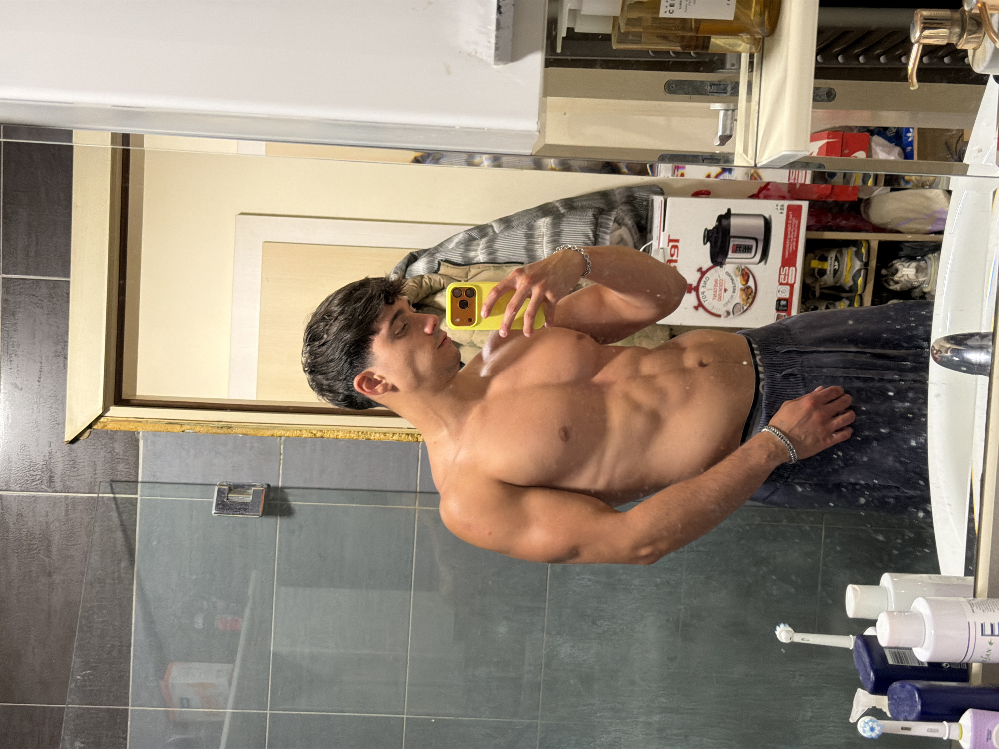A shirtless man with dark hair takes a mirror selfie in a bathroom. He is wearing dark sweatpants and a silver bracelet. His phone is yellow and has multiple camera lenses. The bathroom has tiled walls, a glass shower door, and a mirror that reflects the room. In the background, there are shelves with various items, including toiletries, a pressure cooker box, and shoes. The scene appears to be in a residential bathroom. The location is identified as Sofia, Bulgaria.