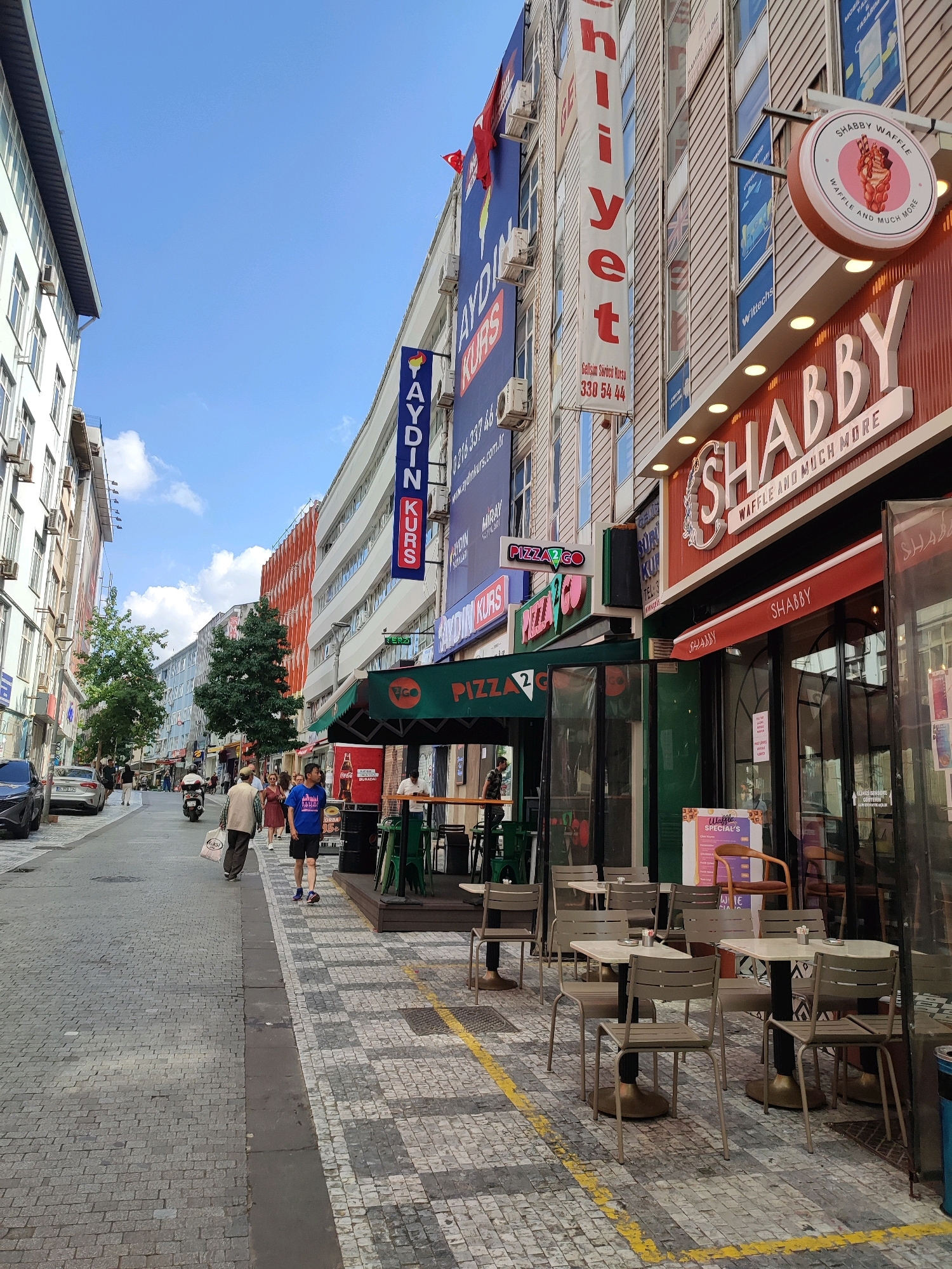 The image shows a street scene in Kadıköy, Turkey. The street is paved with cobblestones, and there are buildings on both sides of the street. On the right side of the street, there is a restaurant called "Shabby Waffle" with outdoor seating. On the left side of the street, there are several shops and businesses, including a sign for "Aydın Kurs" which appears to be a language or education center. Several people are walking on the street, and there are some parked cars and a scooter. The sky is blue with some clouds.