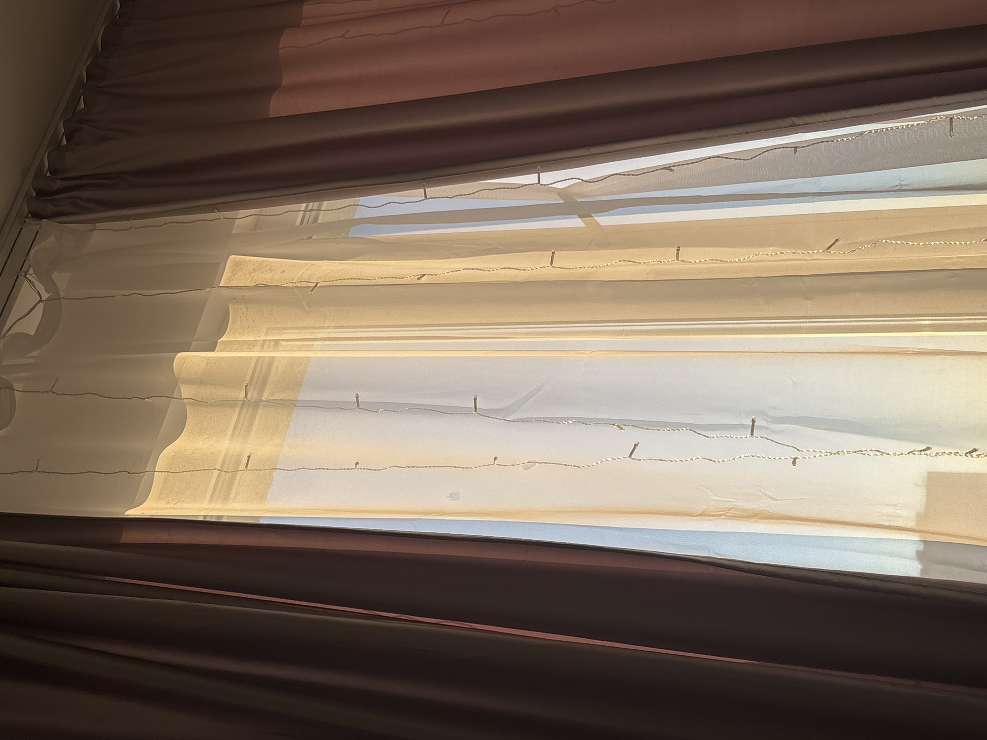 An interior photograph, captured in Unknown, Russia, displays a window dressed with multiple layers of curtains and illuminated by strong natural light. On the left, an opaque dark brown curtain with visible pleats is partially drawn. The central portion of the image features several translucent sheer curtains, predominantly in shades of cream and light beige, heavily backlit by sunlight which imparts a warm, golden glow. Through these sheer layers, subtle blue hues of the sky are perceptible. Vertically suspended across the sheer curtains are multiple strands of decorative string lights, characterized by small, evenly spaced, cylindrical or bead-like illuminators. To the right, a second opaque curtain, dark brown at its upper section, transitions to a distinct muted pink or faded reddish-brown segment below, also partially drawn. A white curtain rod or track system is visible spanning the top of the window frame. No individuals or animate subjects are present, and no specific actions are taking place. The scene depicts a domestic interior.