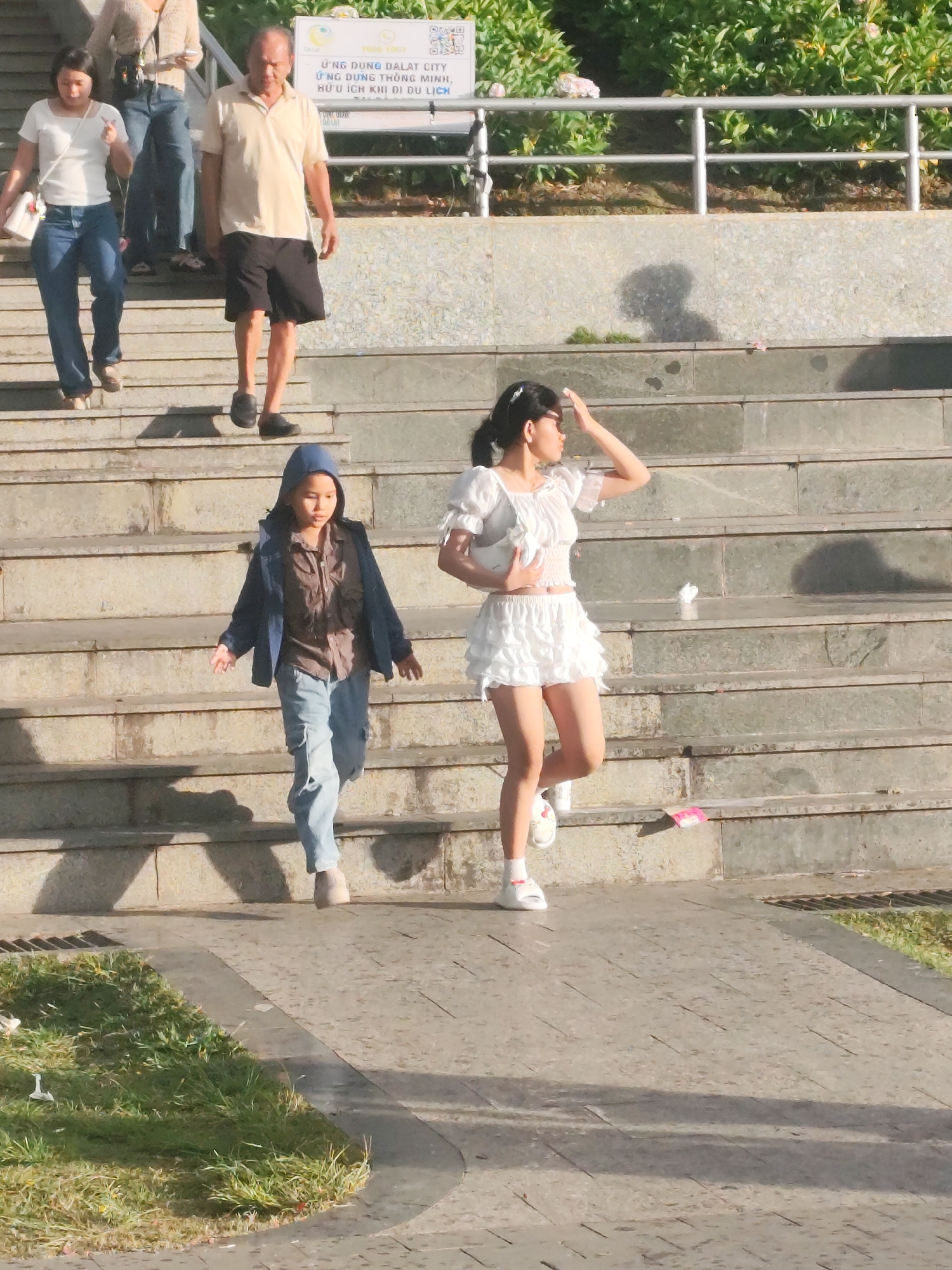The image shows a group of people on a concrete staircase. In the foreground, a young person wearing a blue hooded jacket over a brown shirt and light blue pants walks down the stairs, looking to their right. To the right of the young person, a young woman in a white ruffled top and white ruffled skirt walks down the stairs, holding a white bag and shielding her eyes from the sun with her left hand. Behind them, a man in a light beige polo shirt and dark shorts walks down the stairs. Further up the stairs and to the left, a woman in a white t-shirt and blue jeans is descending, with another person in a light-colored top behind her. The stairs are made of concrete, and there is a railing on the right side of the image. In the background, there is lush green foliage. A sign with Vietnamese text and images is visible on the wall behind the upper part of the stairs. This scene appears to be set in Da Lat, Vietnam, likely in a tourist area or public space with prominent staircases.