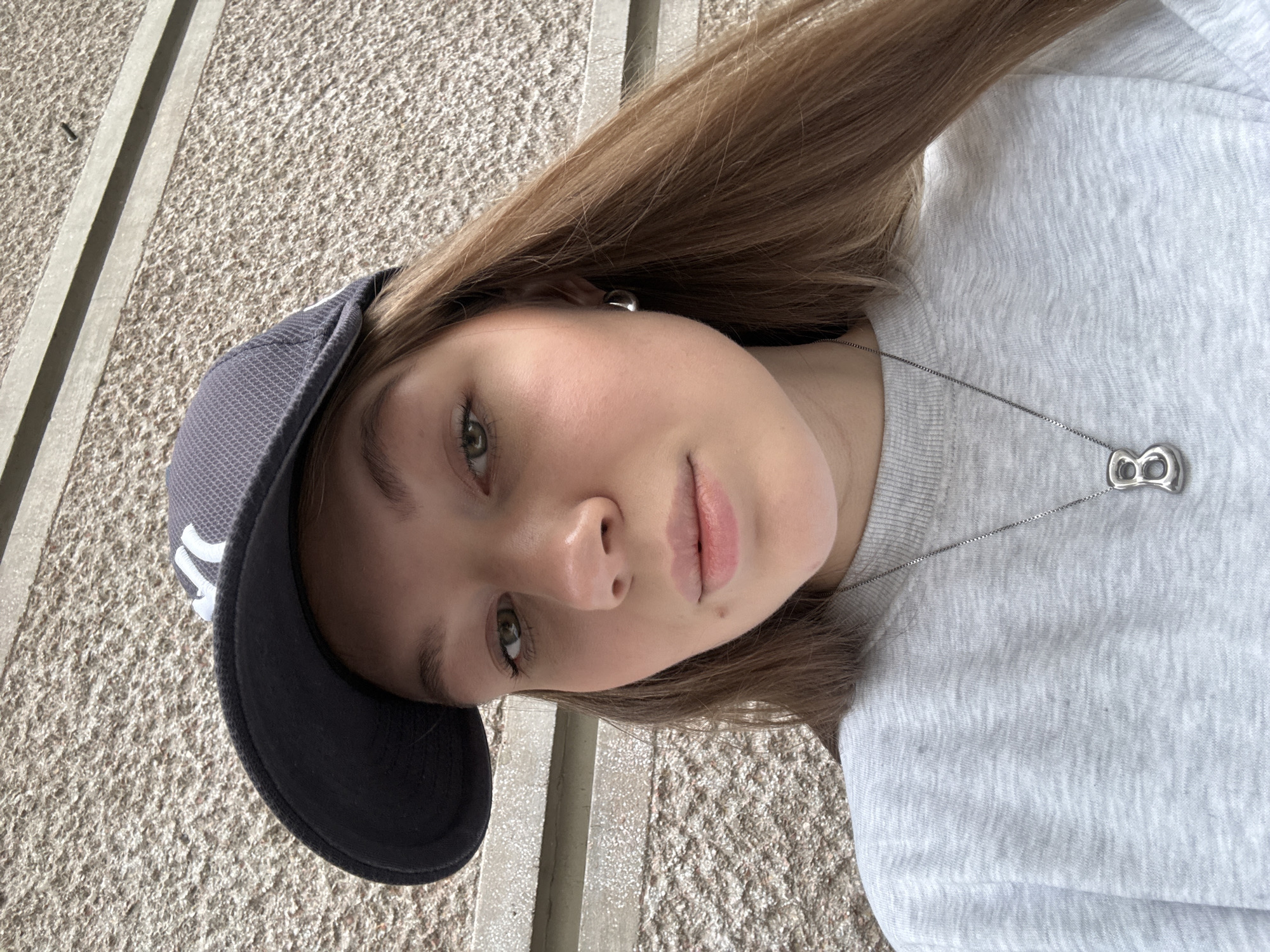 A young female, appearing to be in her late teens or early twenties, is depicted in a close-up, eye-level perspective. She has fair skin, green eyes, and long, straight brown hair that extends over her right shoulder. A small mole is visible below her right nostril. She is wearing a light grey crew-neck sweatshirt and a dark grey baseball cap with a white "NY" logo, positioned slightly askew. A silver-toned hoop earring is visible on her left ear, and a silver-toned chain necklace with a stylized 'B' pendant rests on her chest. The background consists of a textured, light brown or beige stucco wall, consistent with an exterior building facade. A horizontal concrete ledge is visible in the upper right portion of the wall, with a small dark object, possibly a nail or screw, embedded in the wall above it. This scene is located in the city of Belgrade, Serbia.