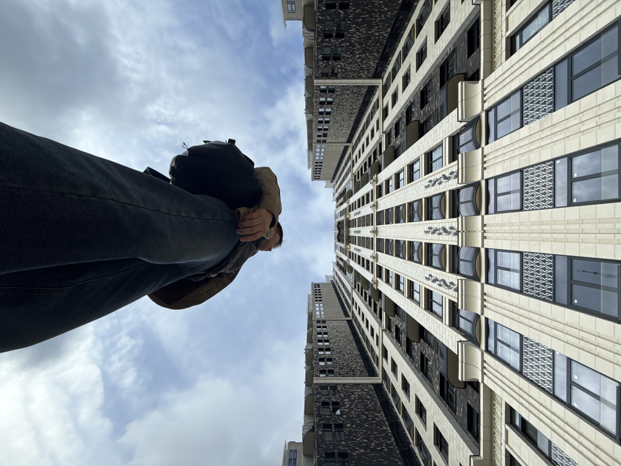 A low-angle shot captures a person standing between two tall buildings in Khimki, Russia. The person, dressed in jeans and a jacket with a black backpack, is looking down. The buildings are modern with detailed facades, consisting of rows of windows. The sky is overcast with scattered clouds. The photograph gives the impression of looking up at the buildings and person.