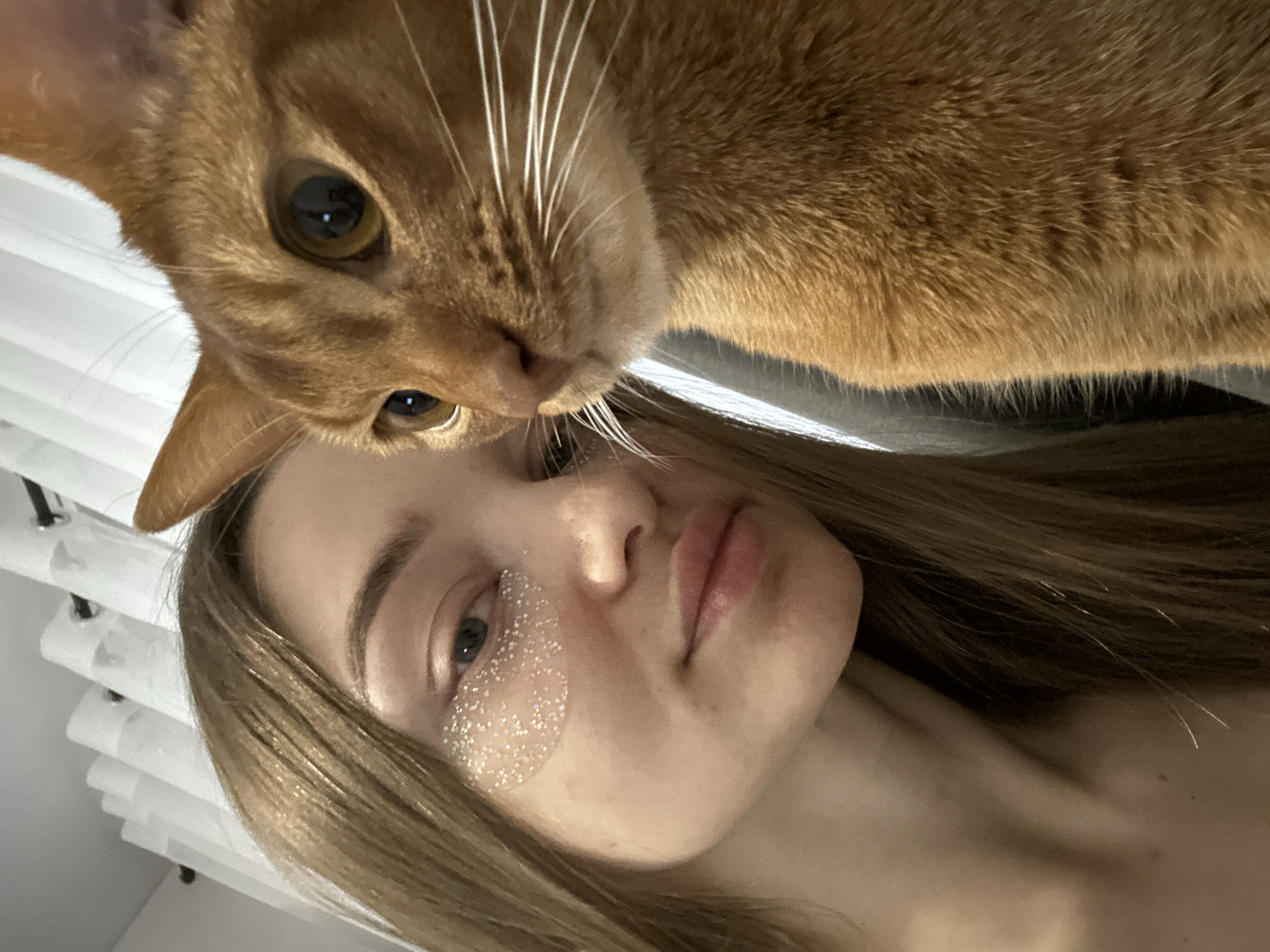 A close-up, slightly tilted photograph shows a woman lying on her back with an orange cat hovering over her face. The woman has fair skin, light brown hair, and appears to be wearing glitter around her left eye. Her eyes are open, and she is looking at the camera. The cat has amber eyes and long white whiskers. The background includes a glimpse of white window blinds. There are no indications that the photo was taken in Kazan, Russia.