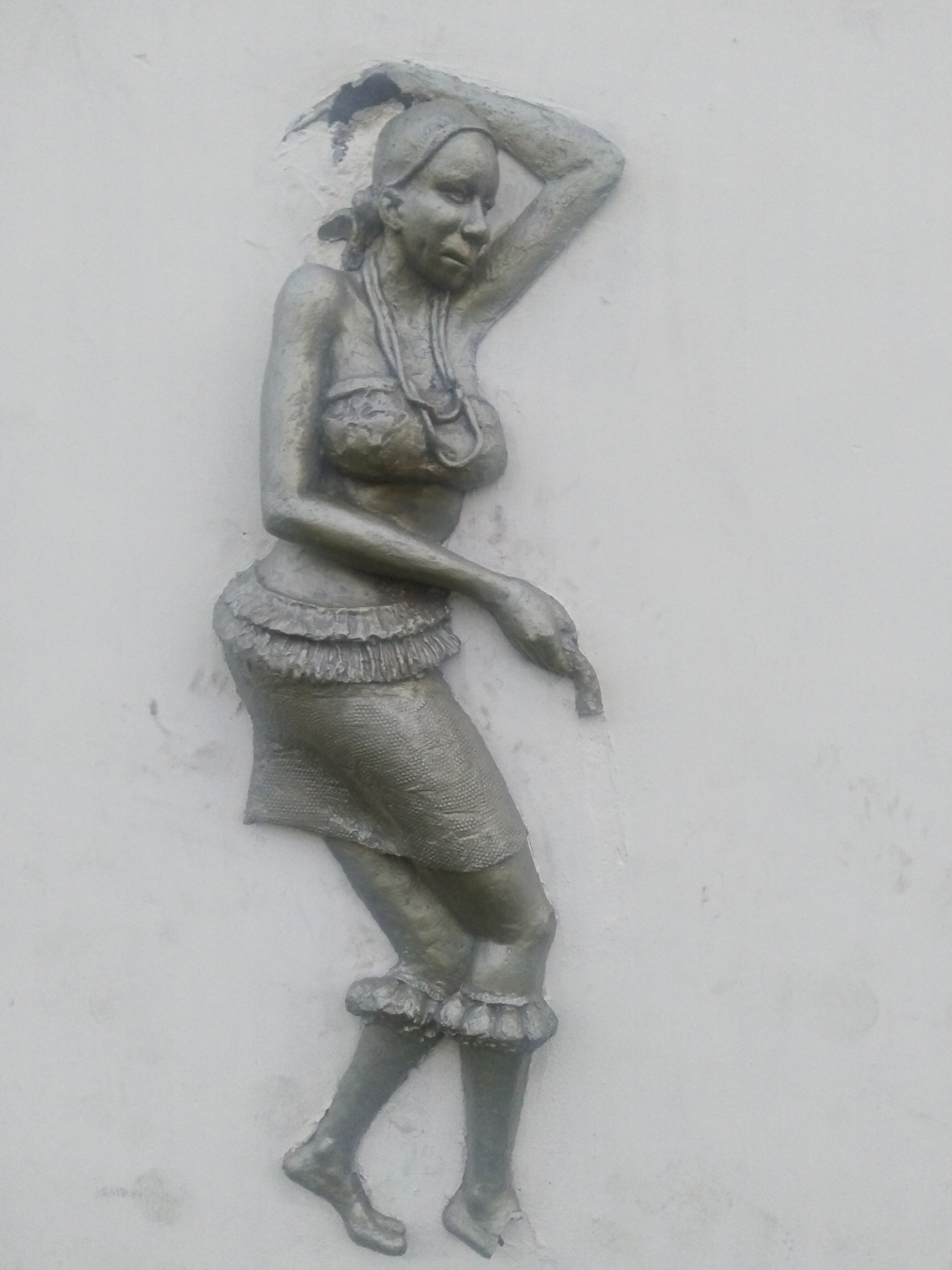 A sculpted relief of a woman is affixed to a white wall. The woman is depicted in motion, possibly dancing. She wears a beaded necklace, a top, and a short skirt with ruffled edges. She has a head covering and her left arm is raised above her head. The sculpture appears to be made of a silver-colored material. It is located in Uyo, Nigeria.