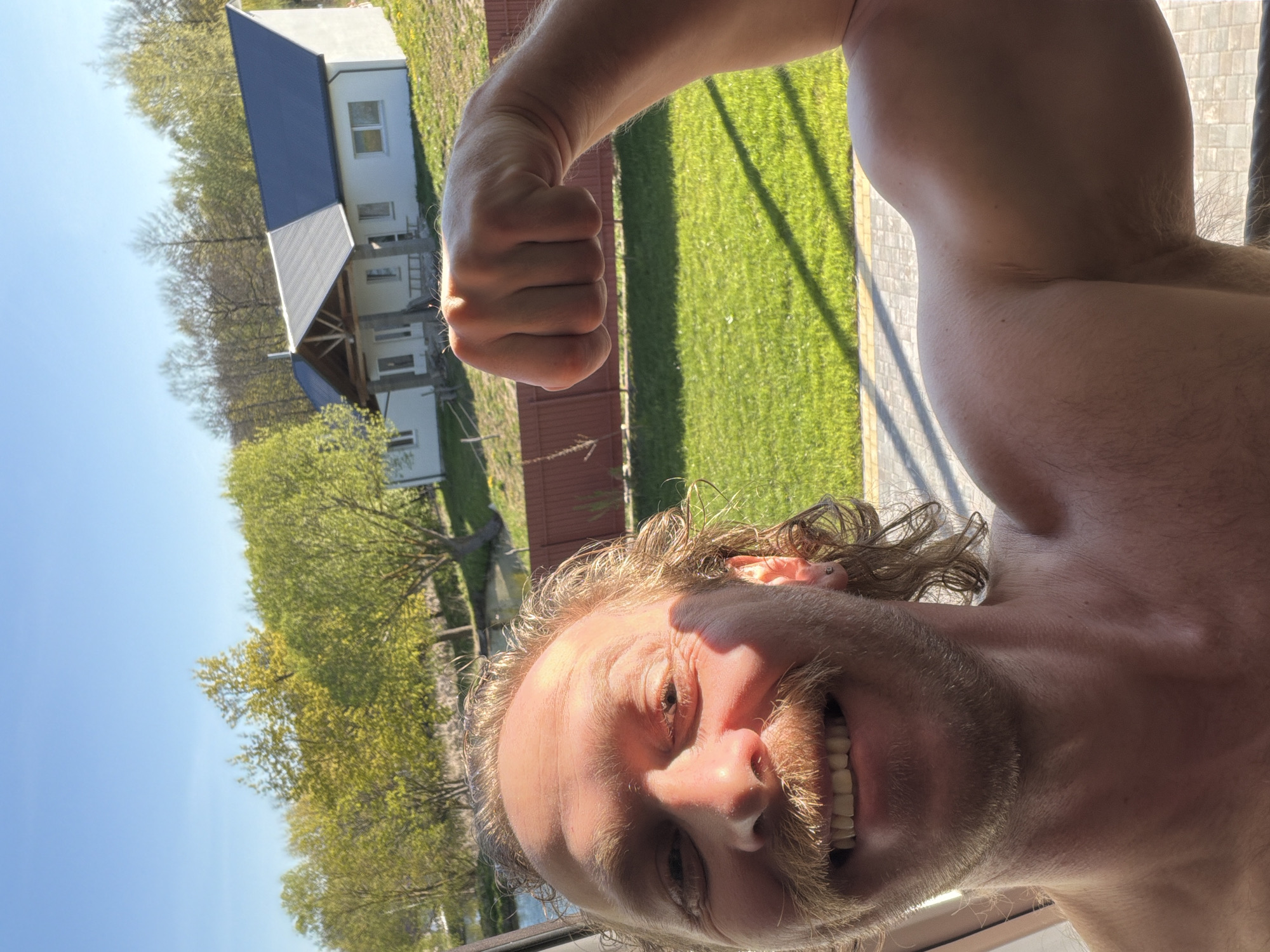 A man with fair skin, curly blonde hair, and a thick mustache smiles into the camera while flexing his right arm and fist. The background features a green lawn, trees with green leaves, and a light-colored building with a dark roof. The image was taken outside, likely on a sunny day. The location is Капцёўскі сельскі Савет, Belarus.