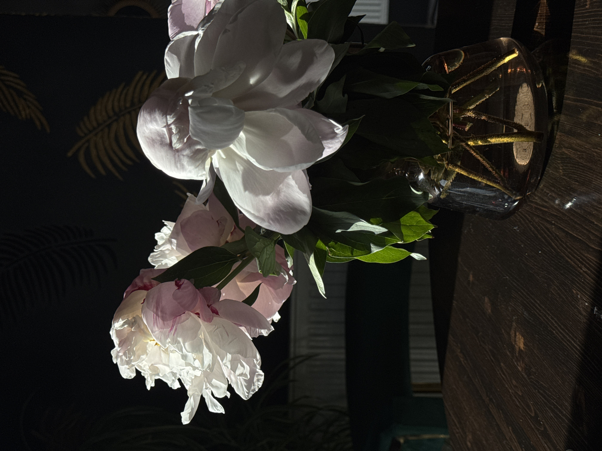 A vase of white and pink flowers sits on a dark wooden surface. The flowers are peonies with green leaves. The vase is clear and contains stems. The background includes a dark wall with gold palm leaf designs. Light is striking the flowers, creating shadows. The image's orientation is vertical, but rotated counterclockwise. The location is potentially Krasnodar, Russia, but there is no explicit evidence for this based on the image alone.