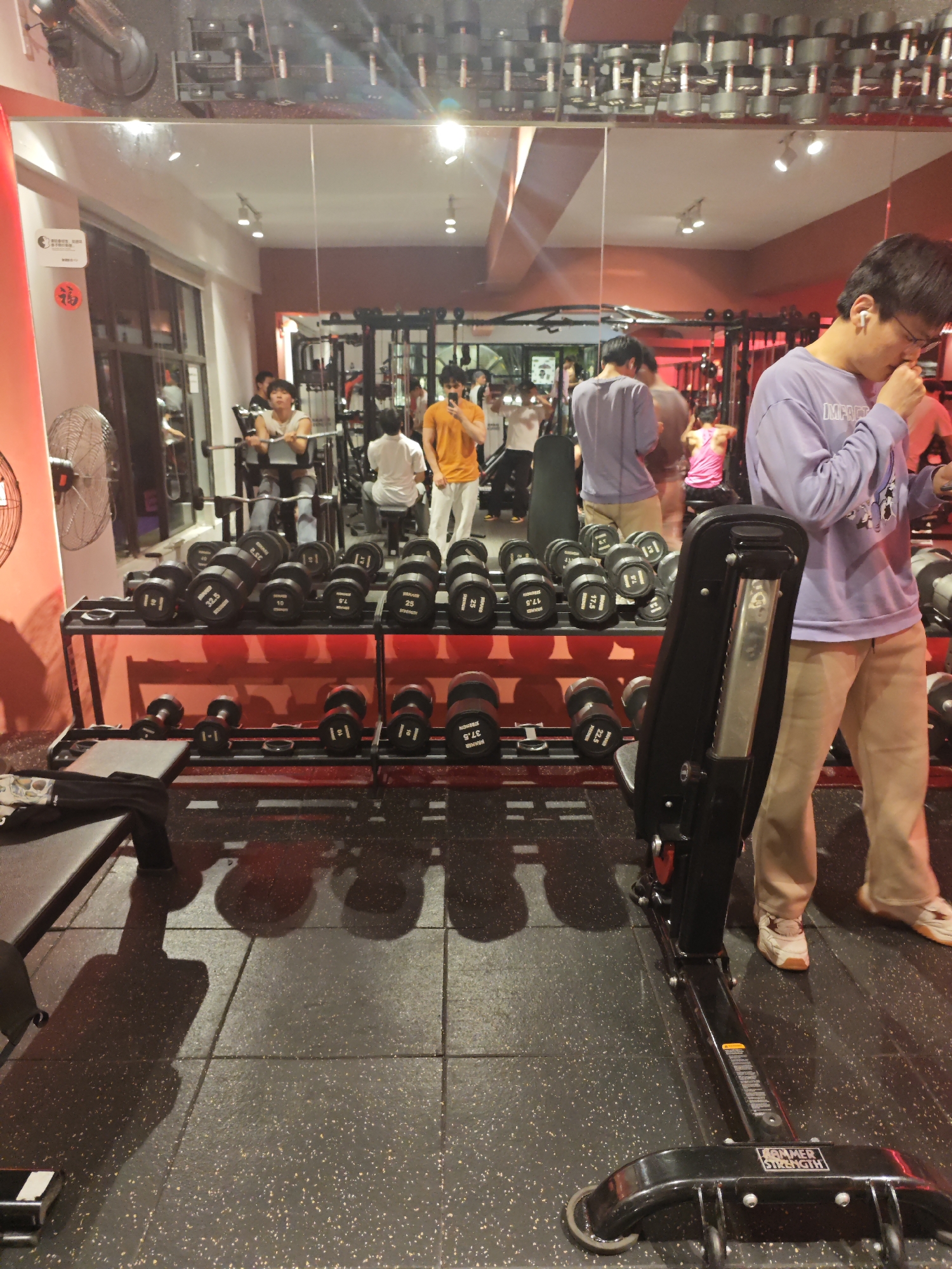 The image shows a gym in the Chongqing High-tech Zone, China. It contains gym equipment such as dumbbells and a Hammer Strength machine. There are multiple people in the gym, including a person wearing a purple shirt and earphones standing near the Hammer Strength machine. Some people are working out and others are standing around. A mirror on the wall reflects the scene, including the gym equipment and people. The lighting is bright and the gym appears well-equipped.