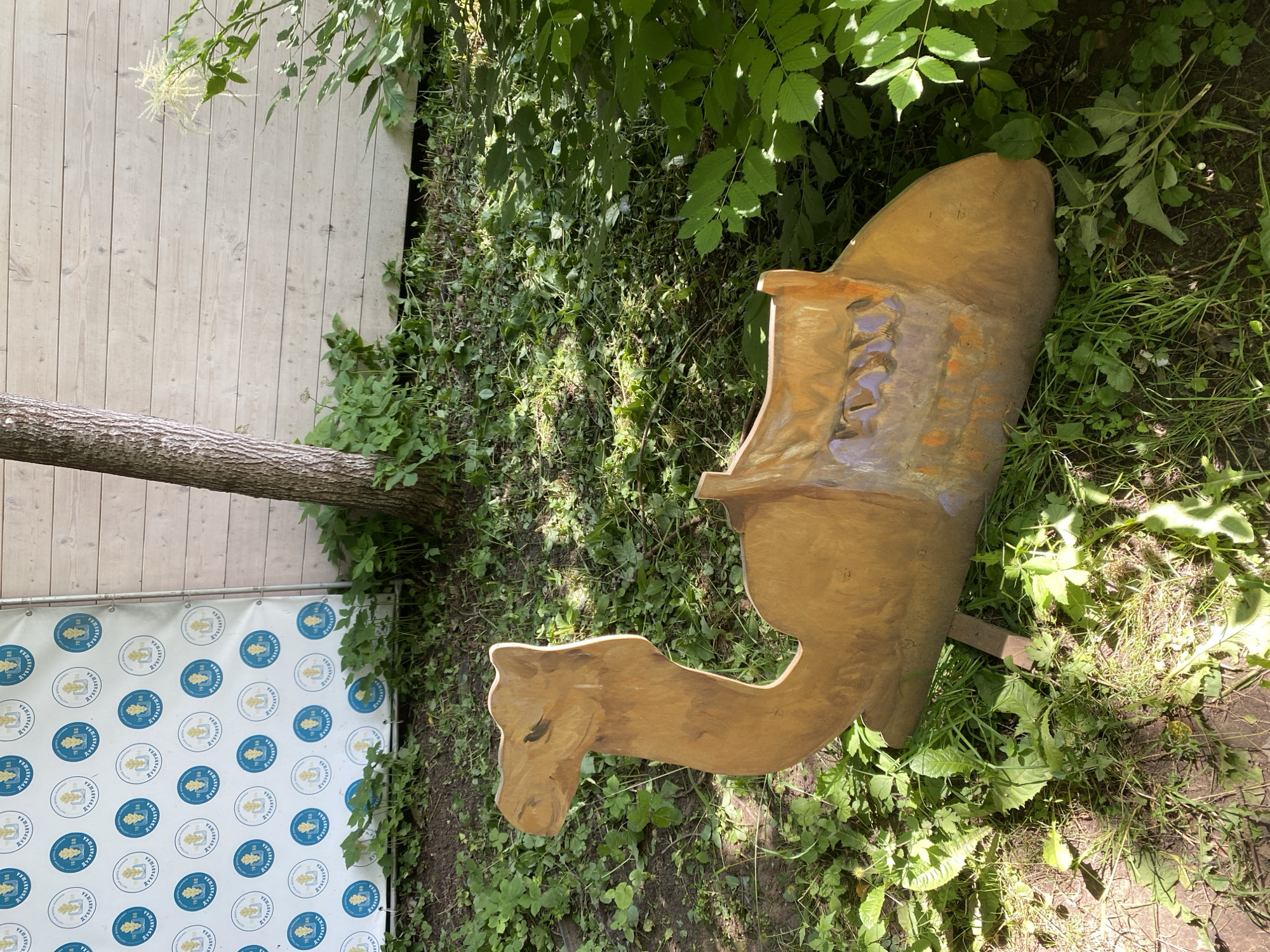 The image shows a wooden camel sculpture placed on the ground amidst lush green vegetation. The sculpture is carved in a sitting position and appears to be weathered. Behind the sculpture, a white wooden fence serves as a backdrop, and a banner with circular logos is affixed to a surface to the left. A tree trunk is visible near the fence. The ground is covered with grass, fallen leaves, and scattered debris. The lighting suggests it is daytime with sunlight filtering through the foliage, creating dappled shadows. The location is specified as Obninsk, Russia. There are no people visible in the image.