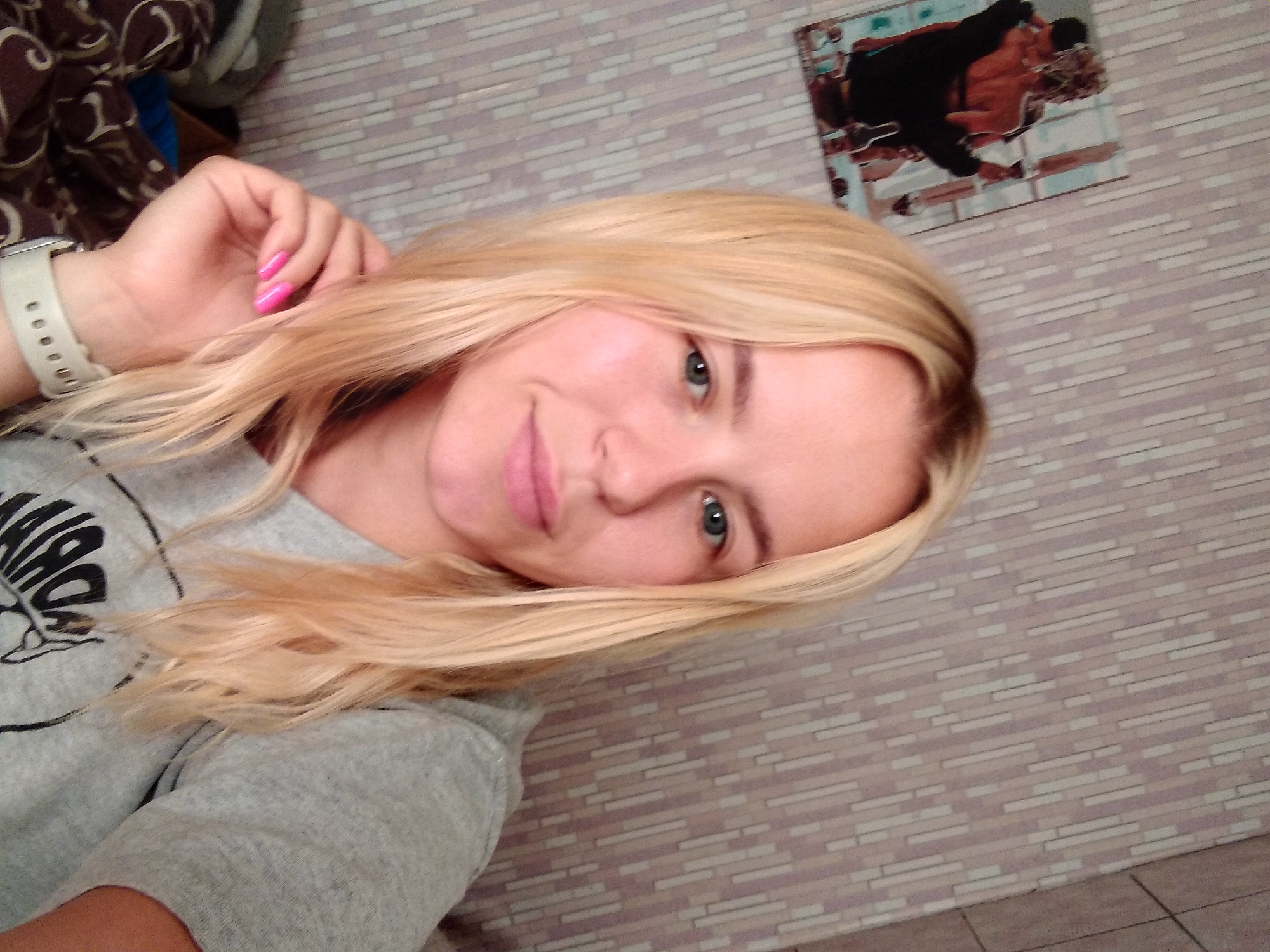 A close-up, waist-up selfie of a young woman with blonde hair and blue eyes is captured indoors. She is wearing a grey t-shirt and has bright pink nail polish on her fingernails. Her left hand is resting on her chin. In the background, there is a wall with a striped wallpaper pattern and a framed picture of a couple embracing. The scene appears to be in a residential setting in Radisne, Ukraine.