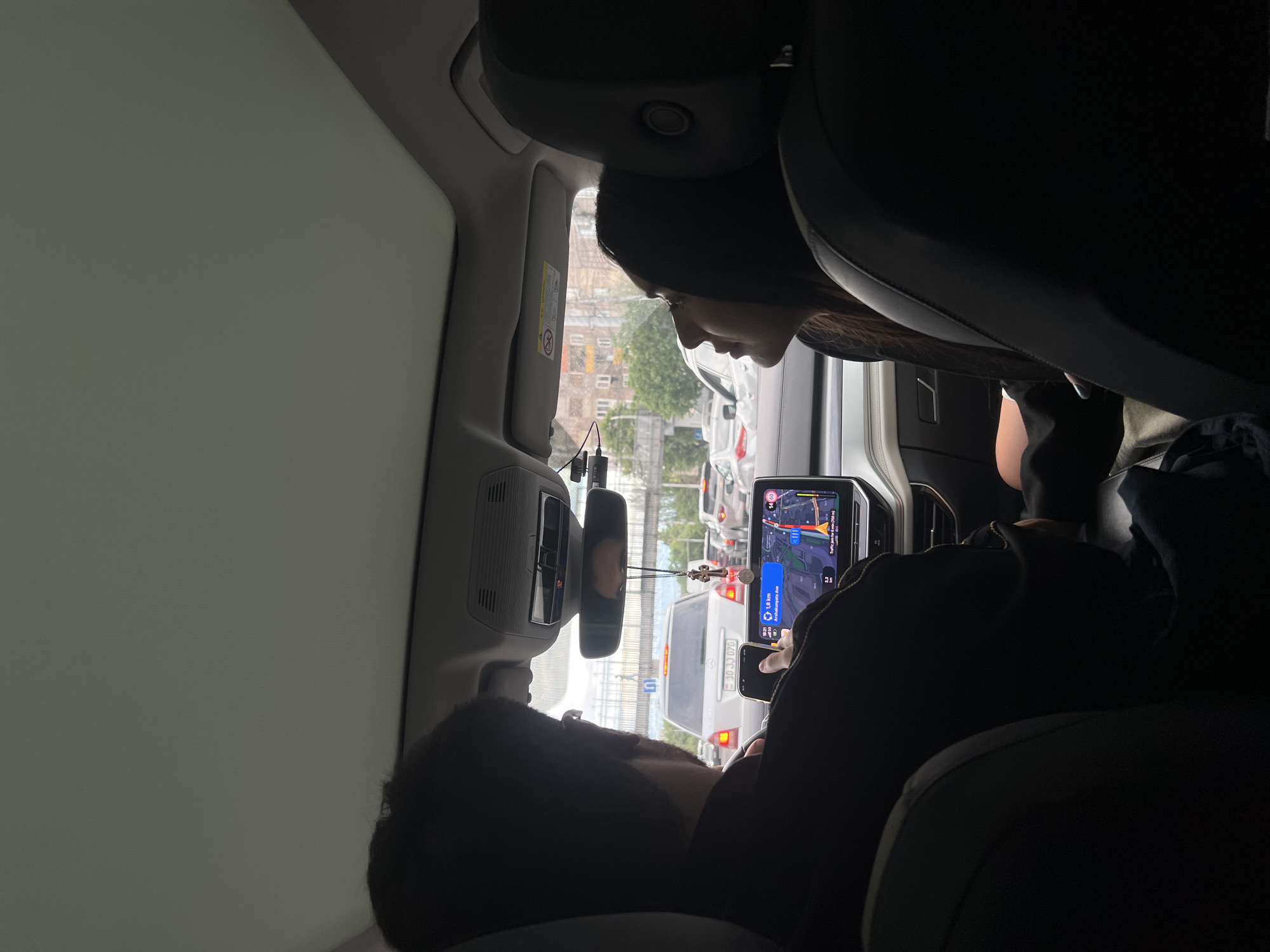 The image shows the interior of a car, likely in Yerevan, Armenia based on the license plate visible on the dashboard phone mount. Traffic is visible outside the vehicle's windows. The rearview mirror is visible above the dashboard. A woman is visible in the front passenger seat. A person is visible in the backseat, possibly reclining.