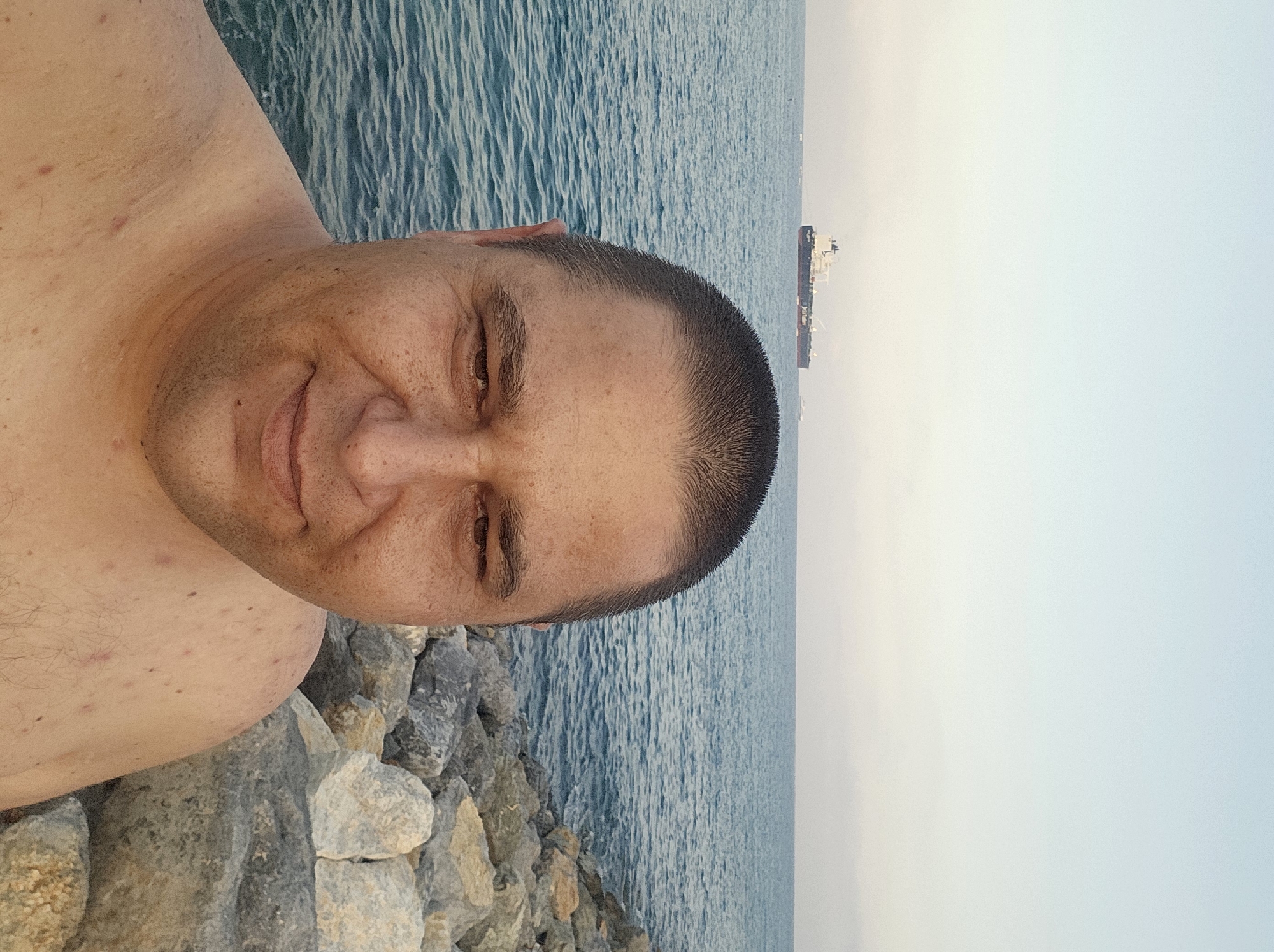 A man is taking a selfie on a rocky shore with the sea and a ship in the background. The man is shirtless and facing the camera. The sea is calm and blue, and the sky is clear. The ship is a large cargo ship sailing in the distance. The rocks are gray and vary in size. The overall scene appears to be a coastal environment. The location is not identifiable as Savona, Italy.