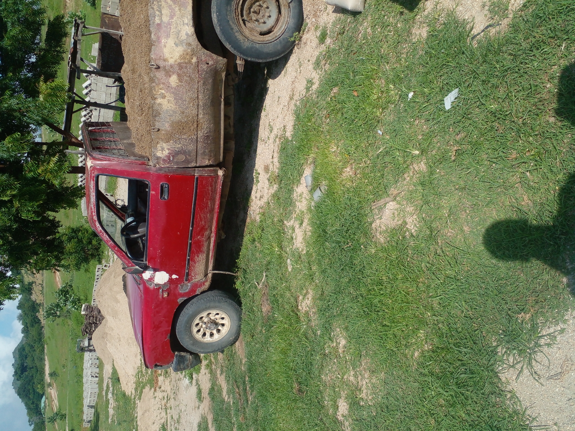 An outdoor, full shot captures a weathered, red pickup truck parked on a patch of dry dirt and grass. The truck is heavily soiled, with a considerable amount of dirt piled in its bed. The vehicle is positioned at an angle, facing towards the left of the frame, with its front half visible and its rear extending out of view. In the background, a lush green hillside is dotted with trees, and in the mid-ground, stacks of concrete blocks and piles of sand are visible, suggesting a construction site. The sky above is a clear blue with scattered white clouds.