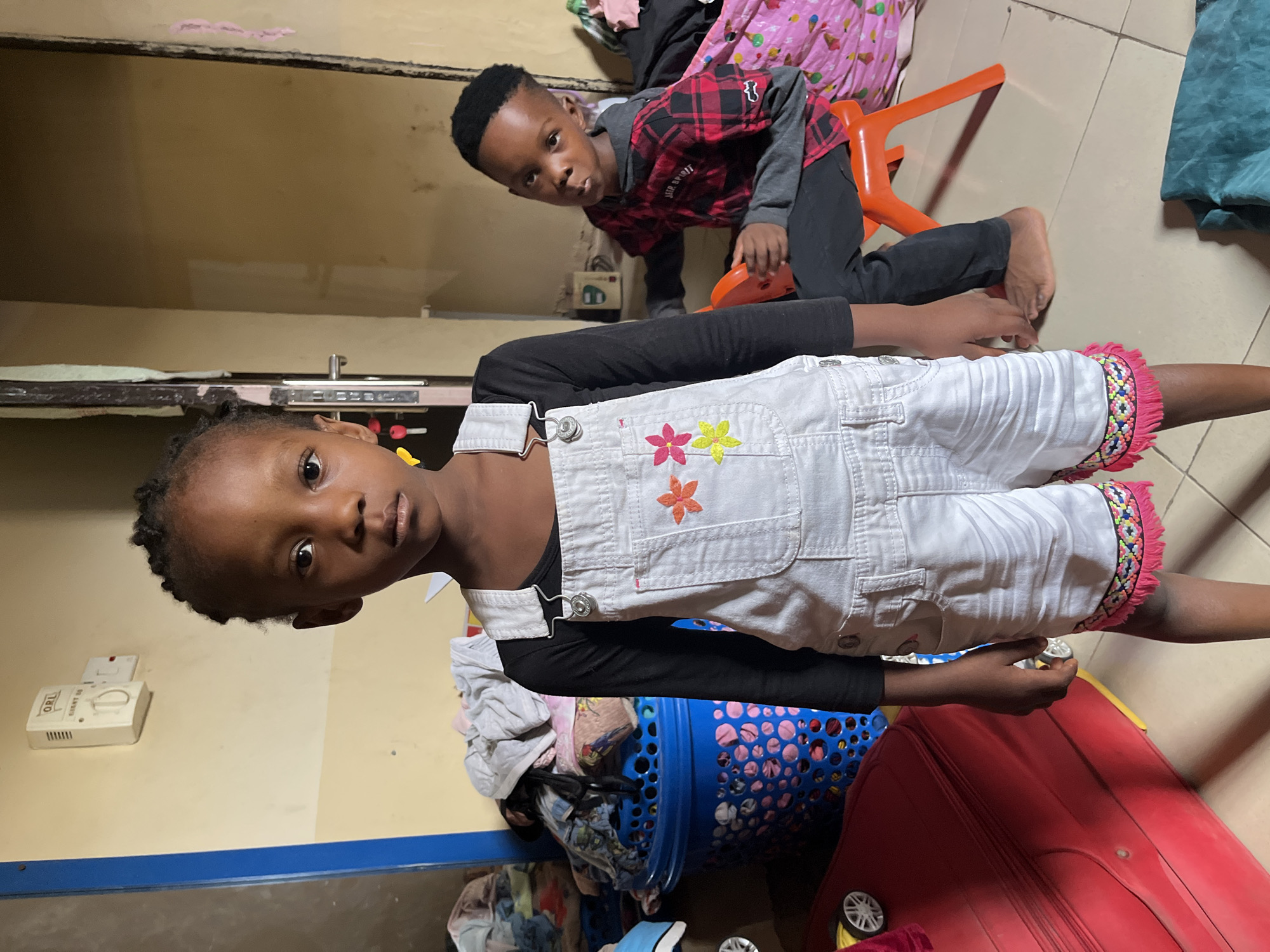 Two children are positioned indoors in Madalla, Nigeria. A girl wearing a black long-sleeve shirt and white overalls with embroidered flowers stands near a blue laundry basket overflowing with clothes, a red suitcase with visible wheels, and a yellow object near her hand. Behind her, a boy in a red and black checkered shirt and black pants leans on an orange plastic chair. The background shows interior walls, an electrical outlet, and various stored items.