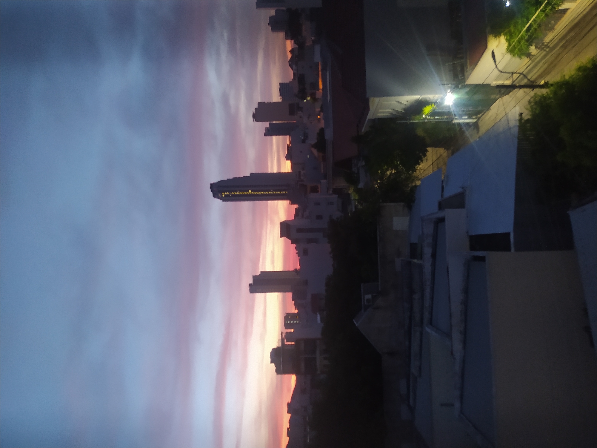 The image is an outdoor, high-angle shot of a city skyline at dusk. The sky is a gradient of colors, transitioning from a deep blue at the top to pink and orange hues near the horizon, indicating the sunset. Silhouetted against the colorful sky are several buildings, including a prominent, tall skyscraper with illuminated windows. In the foreground, the rooftops of lower buildings are visible, some with white surfaces, others appearing darker. Trees and vegetation are scattered throughout the scene, particularly in the mid-ground and obscuring some of the lower buildings. A street with a lamppost and power lines is visible in the lower right portion of the image, illuminated by a bright light. The overall impression is of a city transitioning from day to night.