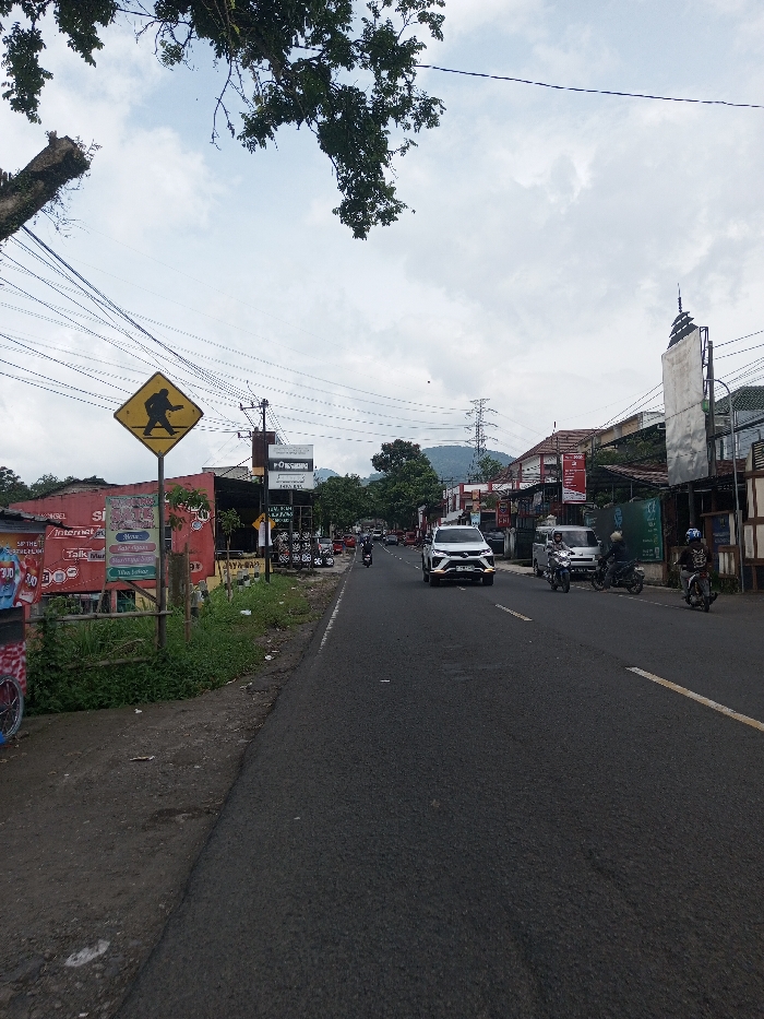 This image captures a street scene in Khajuripada, India, under a partly cloudy sky. The perspective is from a slightly elevated position on the left side of an asphalt road, looking forward along the direction of traffic.
On the immediate left, a dirt shoulder with green grass and weeds borders the asphalt road. A yellow diamond-shaped warning sign stands on a wooden post, displaying a black silhouette of a person in motion, appearing to carry an object over their shoulder. Behind this sign, a structure painted red is partially visible, adorned with multiple commercial banners advertising telecommunication services ("TELKOMSEL," "Internet," "Talk More," "Siam Telkomsel"). A small, partially visible cart with red and white patterns is in the far lower left.
Numerous black power lines and communication cables are strung across the upper portion of the frame, originating from utility poles that line both sides of the road. A large tree branch with green leaves extends from the upper left into the sky.
The asphalt road, marked with a faint yellow line on its right edge, shows active vehicle traffic. A white SUV, possibly a Toyota Fortuner, occupies the center of the road, moving away from the viewer. Behind it, a dark-colored motorcycle follows. Further along the right side of the road, a white van is parked or moving slowly, with at least two motorcycles alongside it; one rider wears a blue helmet. Another motorcycle is visible on the left side of the road in the mid-distance.
Buildings with various commercial signages, including "ForSons" and "Honda," line the right side of the street. A tall, thin, reflective structure, possibly a signpost, rises above the buildings on the right. In the distant background, under the cloudy sky, green hills or mountains are visible, along with a prominent electrical transmission tower.