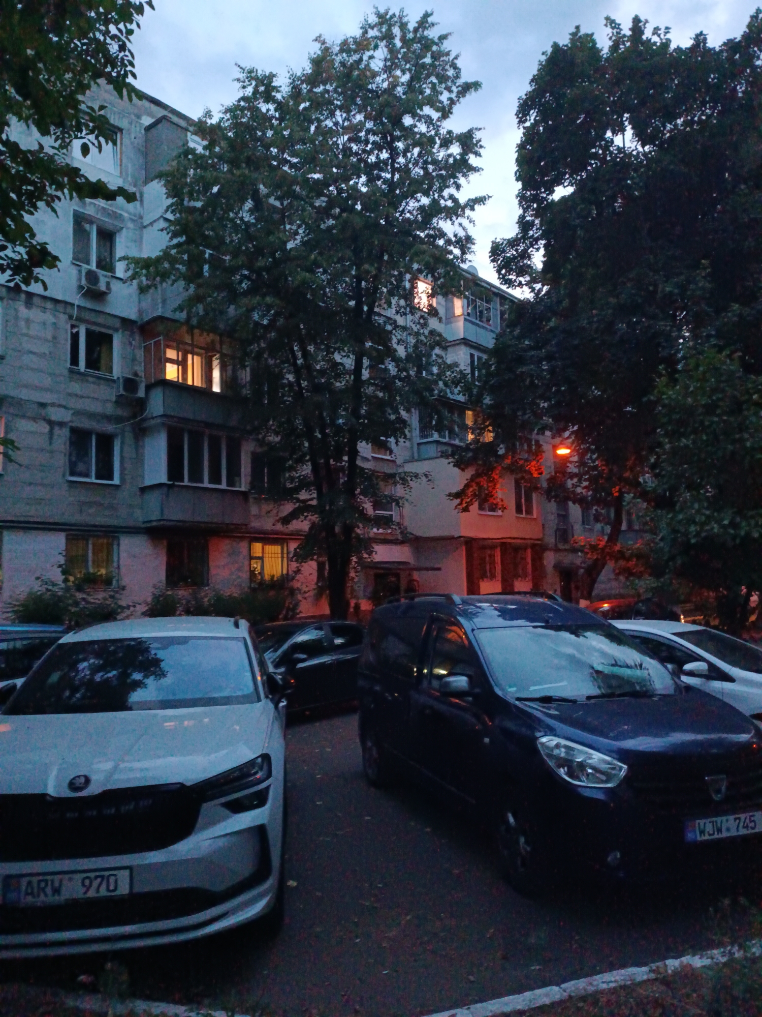 The image depicts a residential street scene in Chișinău, Moldova, during dusk. In the foreground, two cars are parked: a white Skoda SUV with the license plate "ARW 970" on the left, and a dark blue Dacia van with the license plate "WJW 745" on the right. Behind the vehicles is a multi-story apartment building with a grey facade. Several windows are visible, some illuminated from within, suggesting occupancy. Trees with dark green foliage are scattered throughout the scene, partially obscuring the building. A street lamp with a reddish glow is also visible, illuminating some of the tree branches. The sky is overcast with a twilight hue. The overall impression is of a quiet residential area at the end of the day.