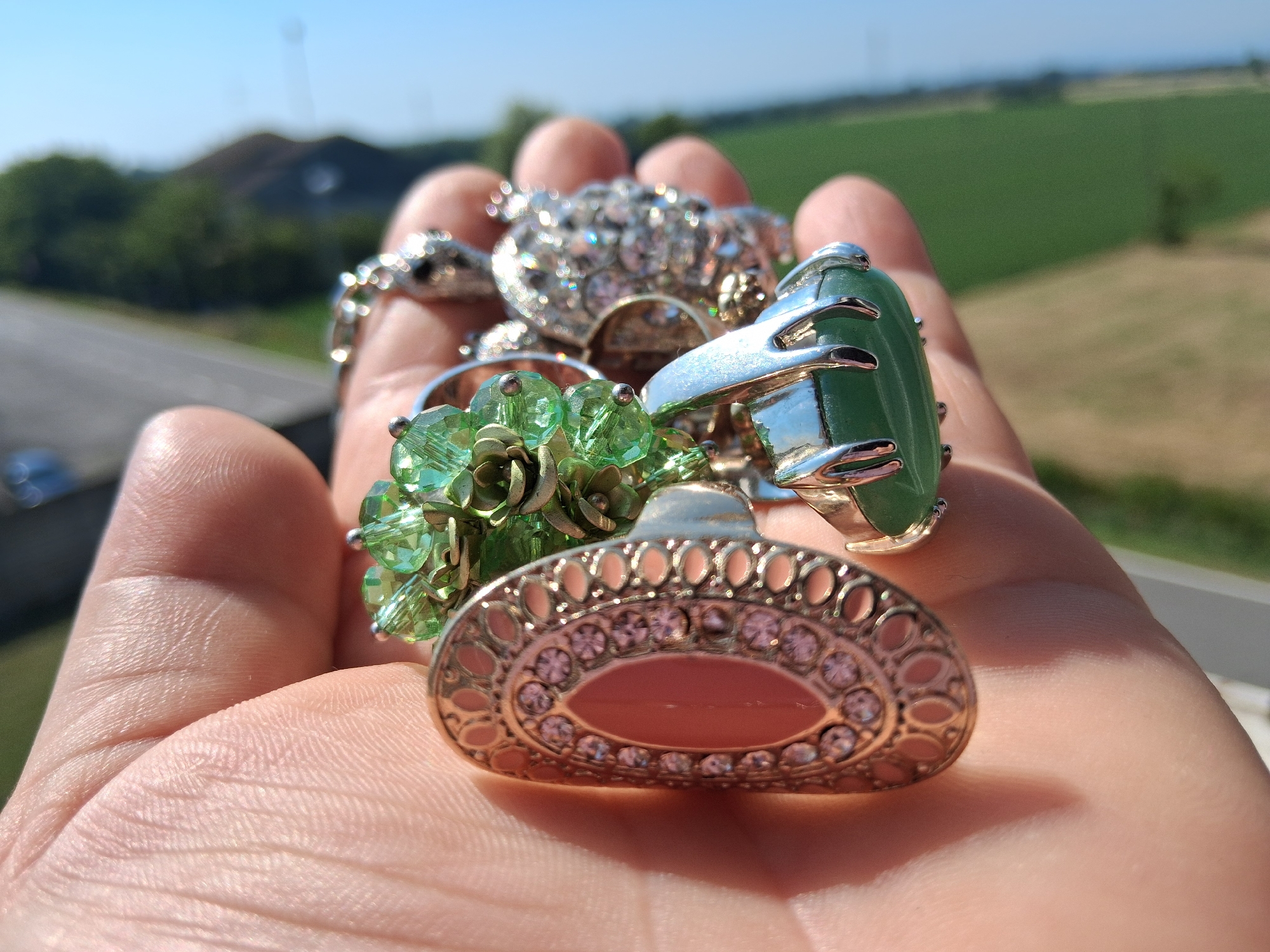 A close-up, eye-level, outdoor shot shows a person's hand holding several rings. The rings are diverse in style and material. In the foreground, a large, oval-shaped ring with a light pink stone is set in an ornate, filigree-like gold-toned metal band adorned with small, clear rhinestones. To the left of this, a cluster of green crystal beads forms a floral design, attached to a silver-toned band. Behind these, a prominent ring features a large, smooth, rectangular green stone, possibly jade, set in a silver-toned metal claw setting. Further back, a snake-shaped ring with numerous clear rhinestones is visible, intertwined with other jewelry. The hand, with visible wrinkles and lines, is gently cupping the rings. The background is blurred, showing a landscape with rolling green fields, some trees, a road, and buildings under a clear blue sky. The lighting suggests a sunny day. No specific landmarks in Colorno, Italy are identifiable.