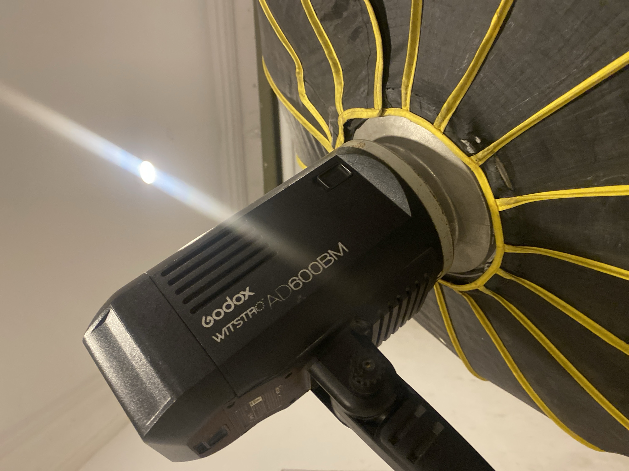 The image presents a close-up, upward-angled view of professional photography equipment, likely in an indoor setting in Badagry, Nigeria. The main subject is a black Godox WITSTRO AD600BM strobe light, clearly identifiable by the visible text "Godox" and "WITSTRO AD600BM" printed on its side. The strobe features vents and a robust mounting bracket.
Attached to the strobe is a black softbox, characterized by prominent yellow internal ribs or baffles that create a striking visual pattern. The softbox appears to be in active use or set up for a photography session, showing minor signs of wear consistent with regular operation.
In the upper left portion of the frame, a bright, intense beam of light emanates from an unidentifiable source, casting a strong, defined glow against a light-colored wall or ceiling. This bright light is a notable detail, hinting at the operational status of the equipment or the presence of another powerful light source in the environment. The overall impression is that of a functional photography setup.
