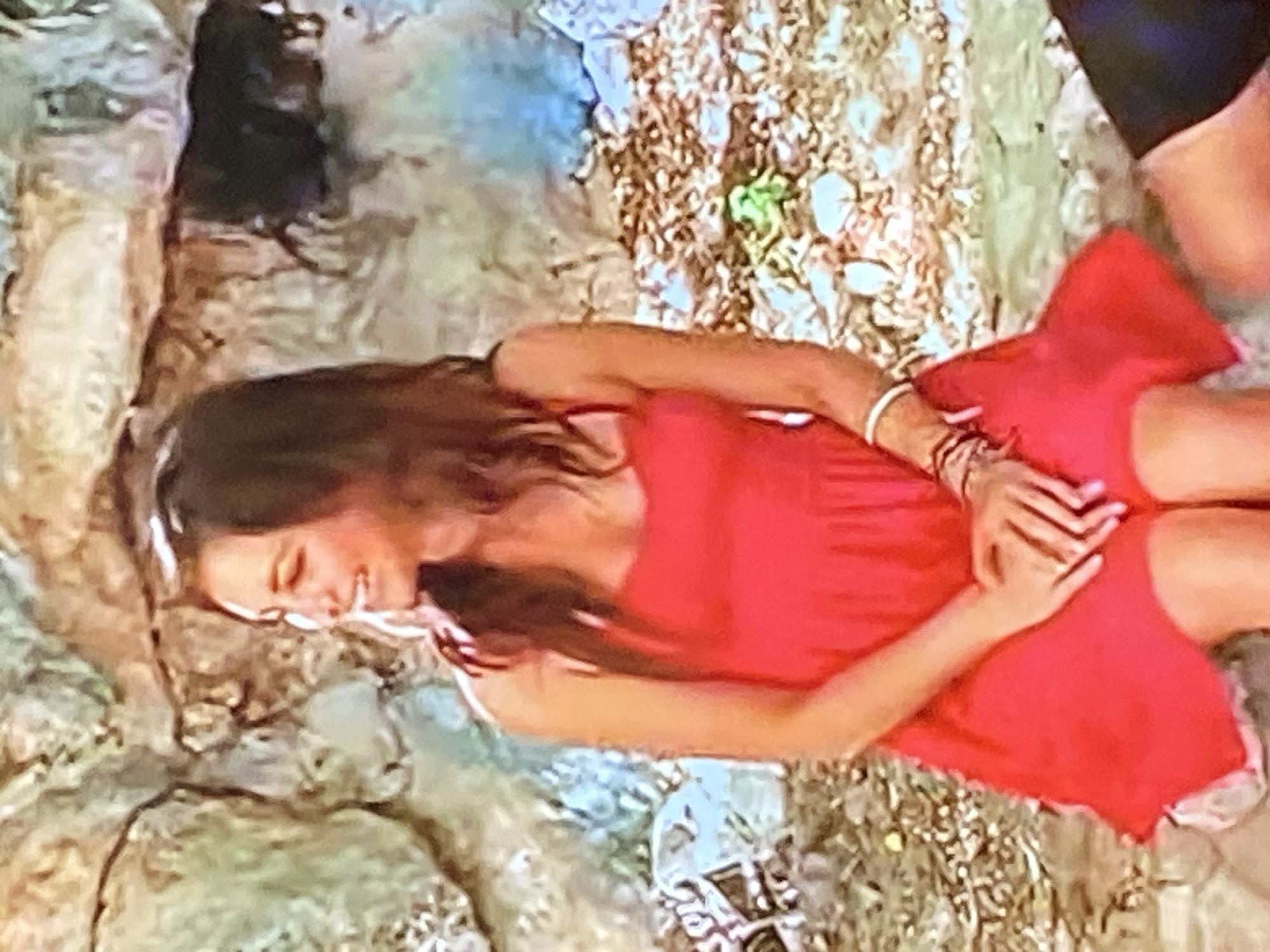 A woman with long, dark hair is sitting in front of a rocky background. She is wearing a short, sleeveless red dress. Her hands are clasped in her lap. She is smiling and looking to her left. The ground around her appears to be covered in small stones and dirt. The background is composed of rough, textured rocks in shades of brown and gray, with some patches of light blue and green. The overall scene appears to be outdoors in a natural or semi-natural environment.