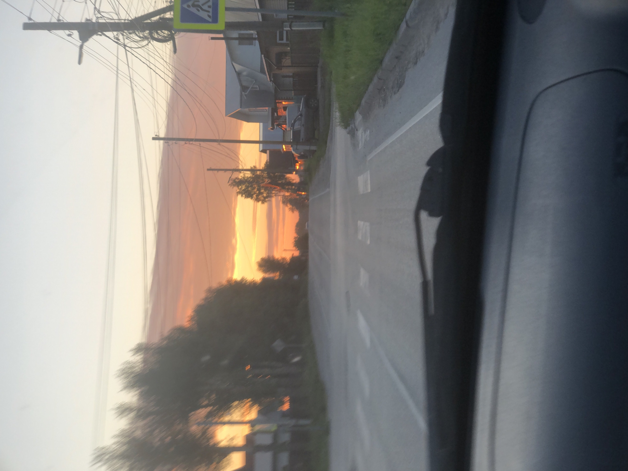 The image depicts a street scene in Solikamsk, Russia, during a sunset. The perspective is from inside a vehicle, looking out at the road and the surrounding environment.
The main subjects are the street, a row of buildings, trees, and utility poles with numerous wires. A pedestrian crossing sign is visible on a utility pole. The sky is dominated by a vibrant sunset, with clouds in shades of orange and yellow, suggesting either dawn or dusk. The sun's rays are casting warm light on the scene.
The road has white lane markings. On the right side of the frame, the interior of the vehicle is partially visible, including a dashboard area and a dark shadow that appears to be cast by the photographer or a passenger.
Notable details include the soft, diffused light typical of sunset, the presence of residential or small commercial buildings lining the street, and the extensive network of overhead electrical or telecommunication wires. The weather appears calm, with no strong indications of wind or precipitation. The overall mood is peaceful, enhanced by the warm colors of the sunset. There is no visible text other than the symbols on the pedestrian crossing sign.