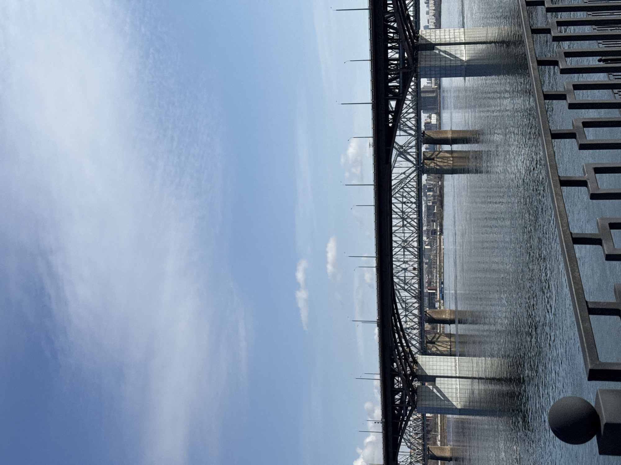 The image shows a bridge spanning a body of water, likely the Yenisey River, in Krasnoyarsk, Russia. The bridge appears to be a steel truss structure with concrete supports. The scene includes a clear sky with wispy clouds. The right side of the image captures part of a waterfront area with decorative railings. The water reflects the bridge and sky. No people are visible.