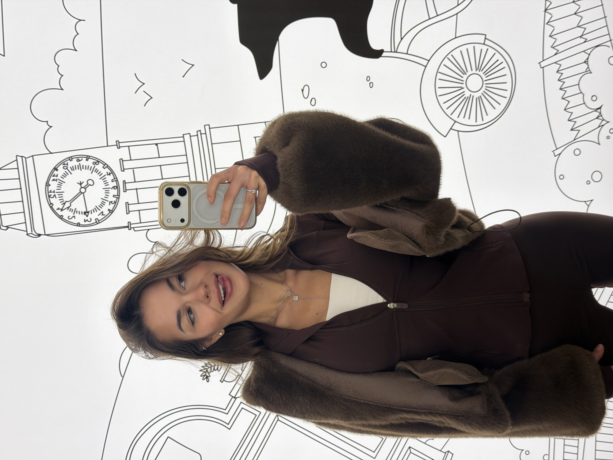 A woman is taking a selfie in front of a mural depicting London landmarks, including Big Ben and a double-decker bus. She is wearing a brown zip-up jacket with fur trim and brown leggings. She holds a smartphone with a light-colored case. The mural consists of black line drawings on a white background.