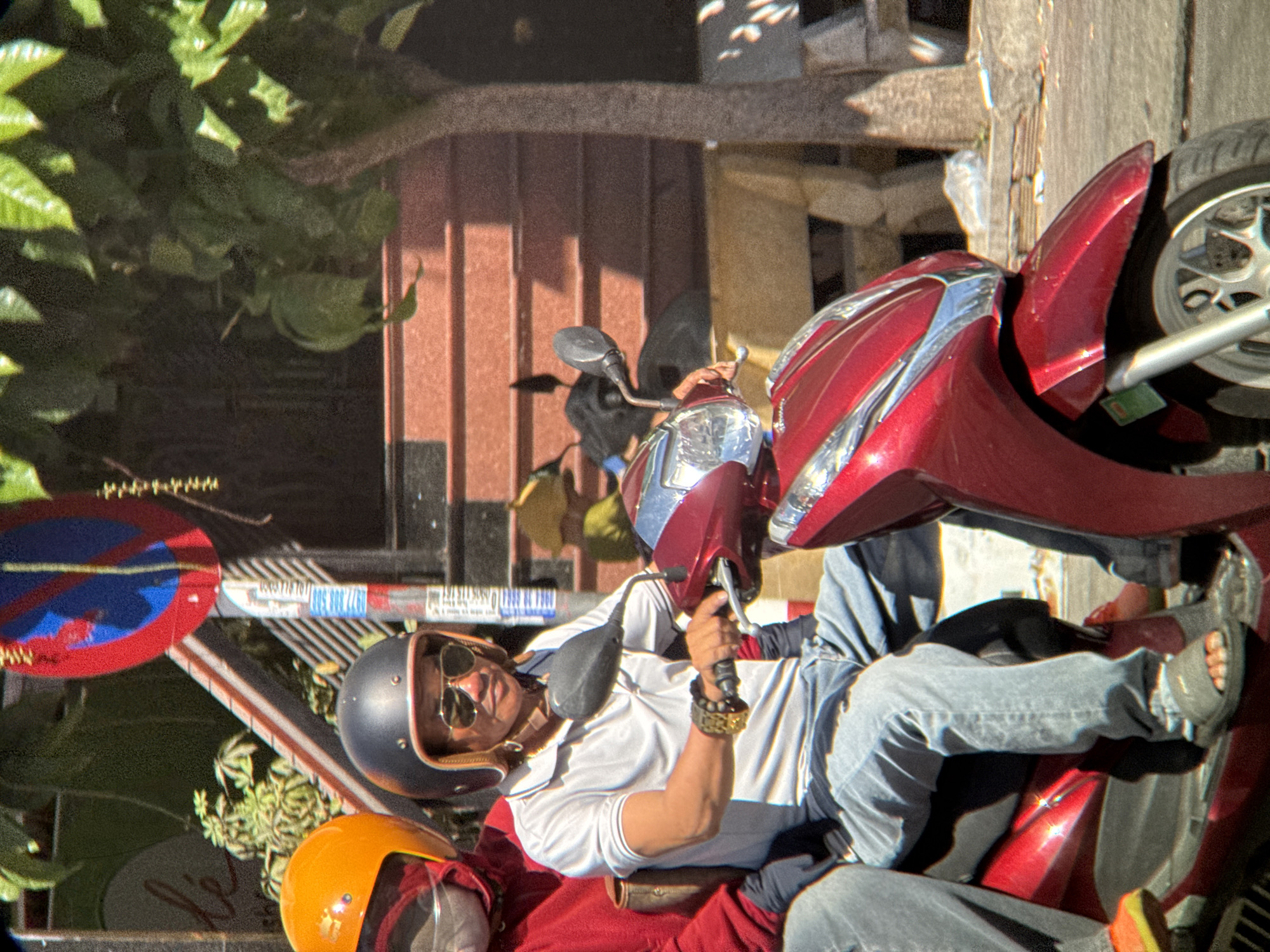 A man and woman are riding on a red scooter on a street in Nha Trang, Vietnam. The man, in the front, is wearing a white polo shirt, blue jeans, sunglasses, and a black helmet. He has dark hair and is looking forward. The woman, behind him, is wearing a red shirt and an orange helmet. She is partially obscured by the man. The scooter is a modern, sporty model. In the background, there is a building with red siding, a tree, and a traffic sign with red and blue markings. The scene appears to be a typical urban street in Vietnam, with bright sunlight and a sense of movement.
