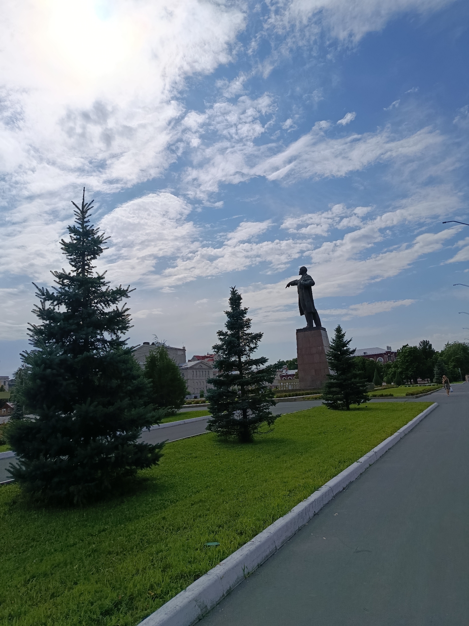 This image captures a scene in Saratov, Russia, featuring a large statue of Vladimir Lenin prominently placed on a stone pedestal. The statue is situated in a park-like setting with well-maintained green grass and several evergreen trees, including a large spruce in the foreground on the left. In the background, there are buildings that appear to be part of the city's architecture. The sky above is blue with scattered white clouds, and the sun is partially visible. A paved walkway runs along the right side of the image, and a person is walking on it in the distance. The overall impression is a public space in a city, likely a square or a park, with a historical monument as its focal point.