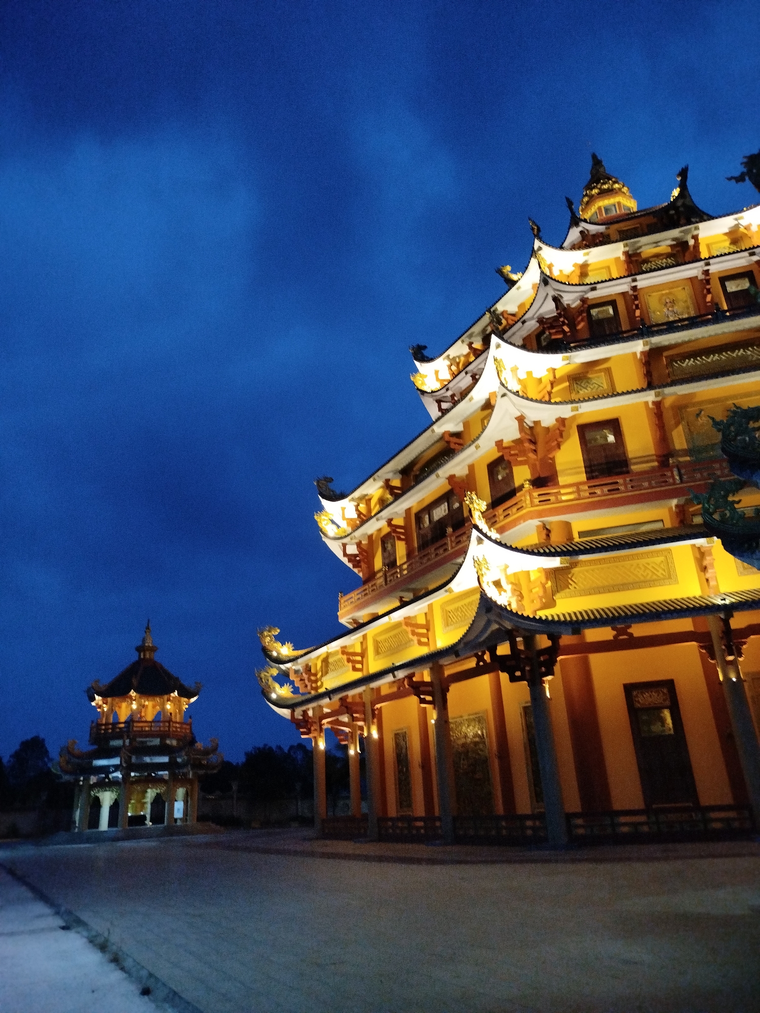 A low-angle shot captures two pagodas illuminated against a dark blue evening sky in Phú An, Vietnam. The larger, multi-tiered pagoda on the right is brightly lit in yellow and white. Its architecture includes dark-tiled, ornate roofs with upturned corners and dragon-like ornaments. The smaller pagoda on the left, in the background, is similarly lit, but less intensely. The ground in the foreground is a concrete or stone surface, only partially illuminated. Trees are visible in the background between the pagodas.