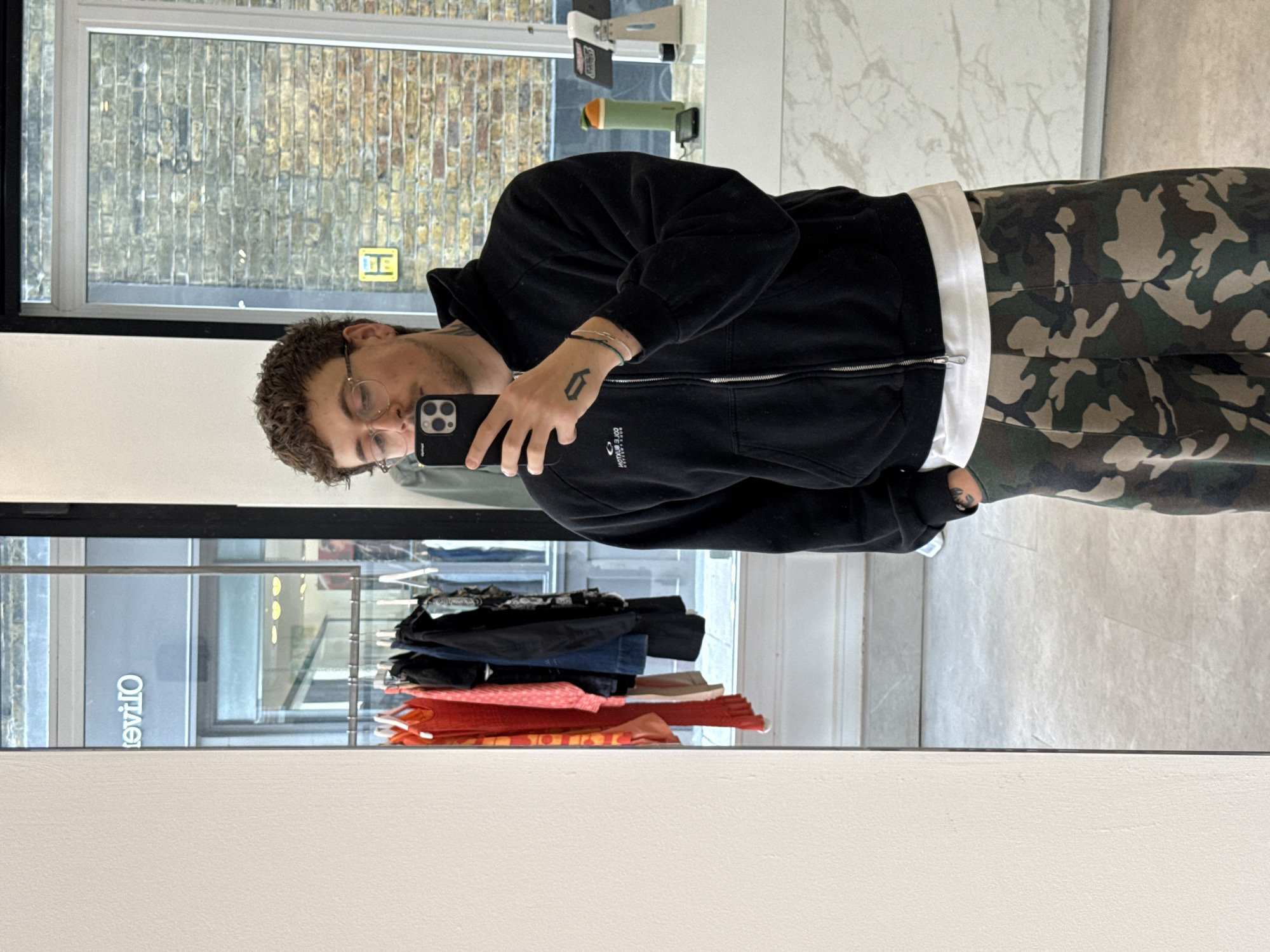 A person with curly brown hair and glasses is taking a mirror selfie in a shop. They are wearing a black zipped hoodie, a white t-shirt, and camouflage trousers. The reflection in the mirror reveals clothing racks with folded garments. Outside the window, a brick building typical of London is visible. The location appears to be a retail store in London, United Kingdom.