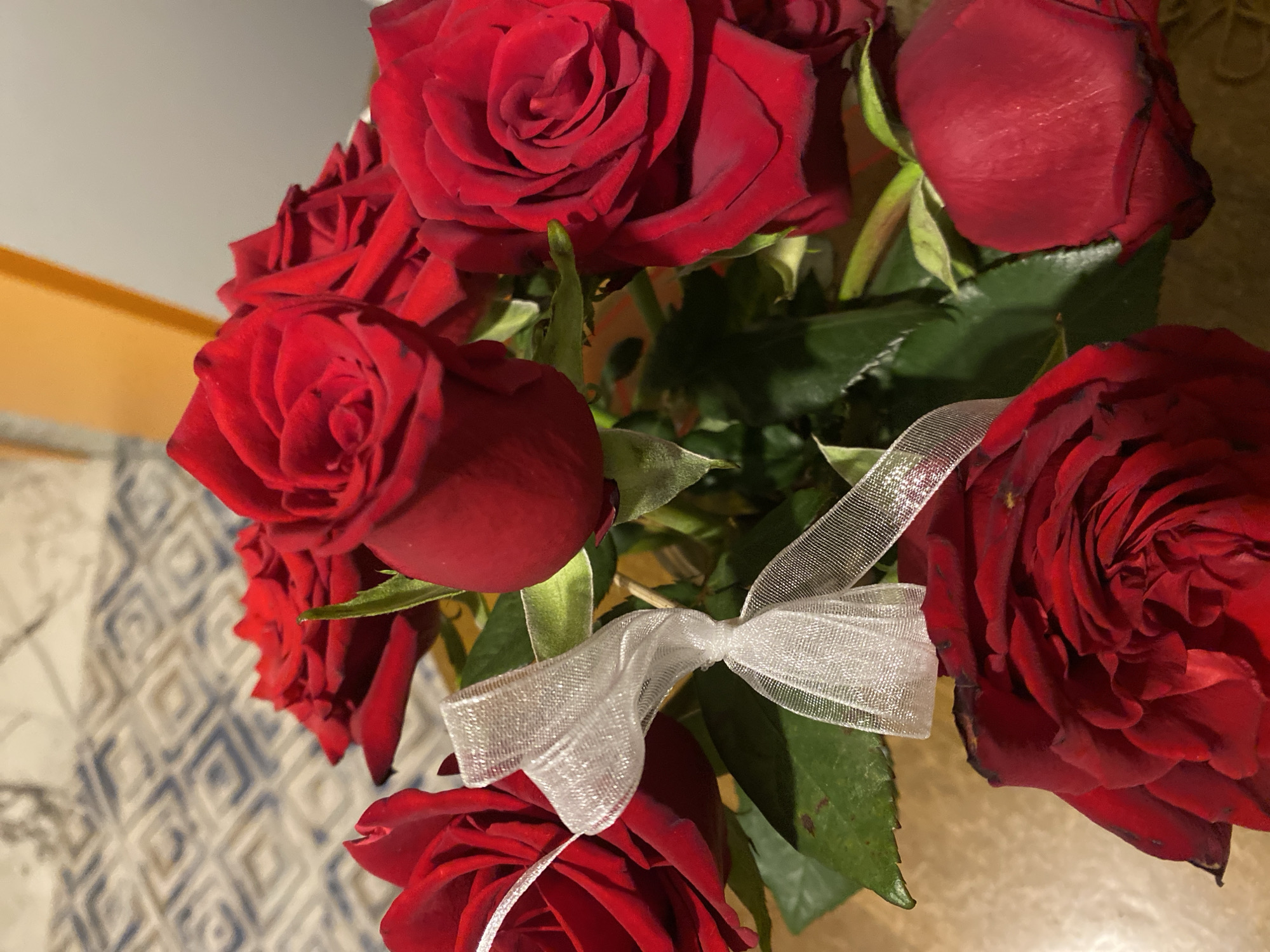 A close-up, high-angle shot depicts a bouquet of deep red roses adorned with a delicate, sheer white ribbon tied into a bow. The roses are in varying stages of bloom, some tightly closed buds and others fully open. Green leaves intermingle with the flowers, providing contrast. The background is slightly out of focus, revealing a surface with a patterned design. There's no evidence suggesting the image was taken in Alanya, Turkey or any other location; there are no location specific landmarks or identifiers.