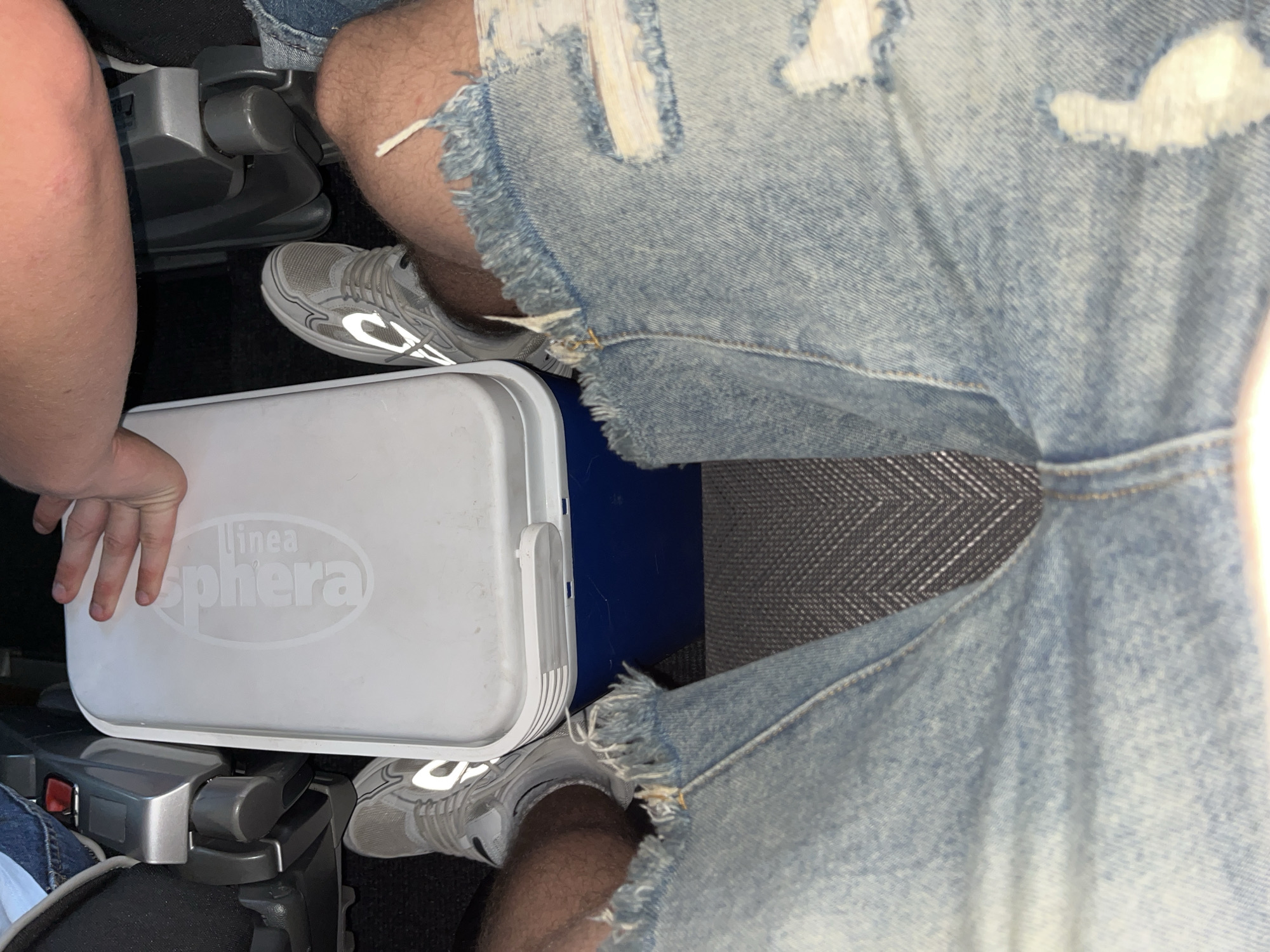 The image shows a person seated, possibly on an airplane or bus, with a white "linea Sphaera" cooler placed between their legs. They are wearing light blue, ripped denim shorts and gray sneakers with reflective detailing. The seat has a gray patterned fabric. The exact location in Ca' Savio, Italy, cannot be determined from the image alone.